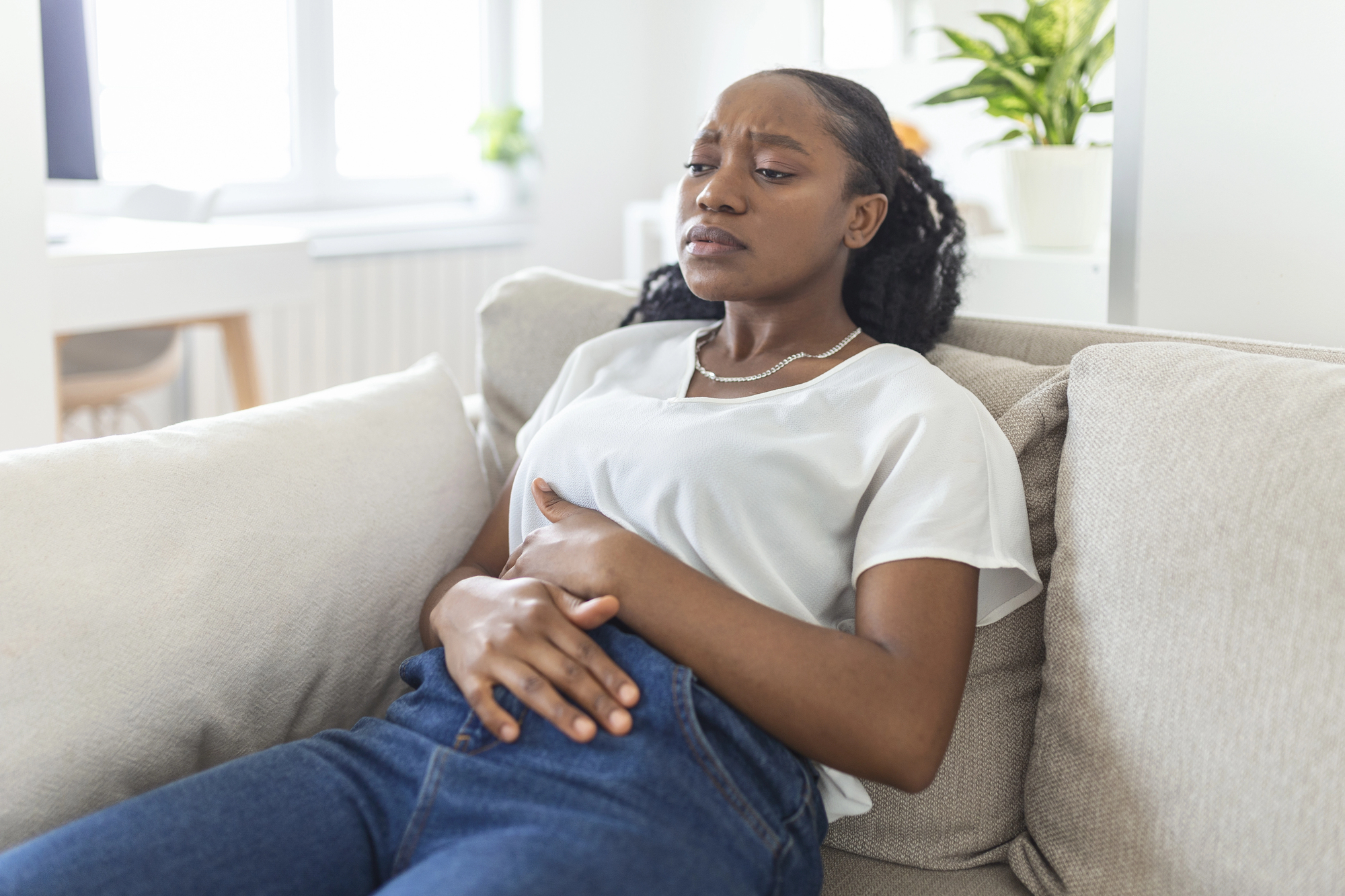 A woman sitting on a sofa holds her stomach with both hands, appearing uncomfortable. She is wearing a white shirt and blue jeans. In the background, there's a bright room with a window and a potted plant.