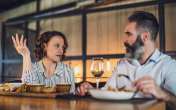 A woman and a man are sitting at a table in a restaurant, engaged in conversation. The woman gestures with her hand while the man focuses on his meal. There are plates of food and a glass of wine in front of them.