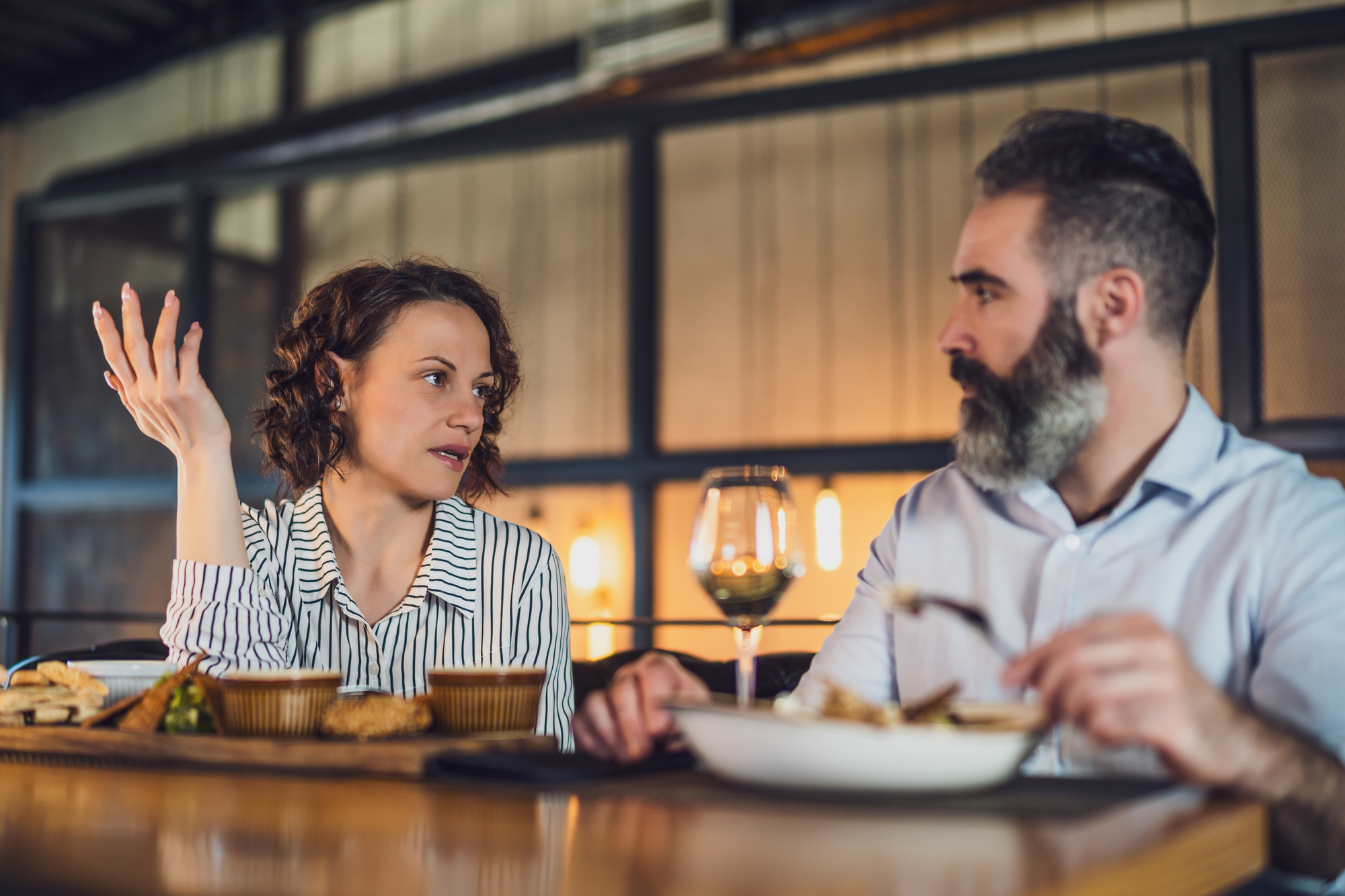 A woman and a man are sitting at a table in a restaurant, engaged in conversation. The woman gestures with her hand while the man focuses on his meal. There are plates of food and a glass of wine in front of them.