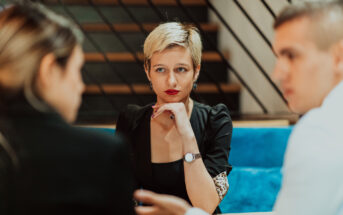 Three people are engaged in a serious conversation in a modern setting. A woman with short blond hair sits in the middle, listening intently to the two people beside her. The background features a staircase with diagonal railing.