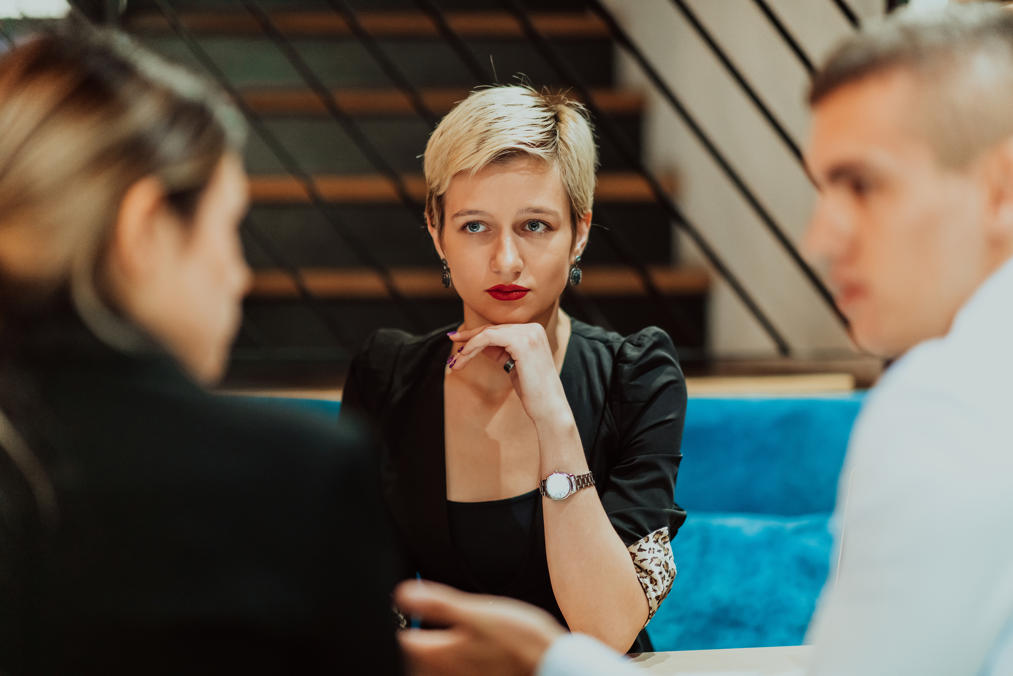 Three people are engaged in a serious conversation in a modern setting. A woman with short blond hair sits in the middle, listening intently to the two people beside her. The background features a staircase with diagonal railing.