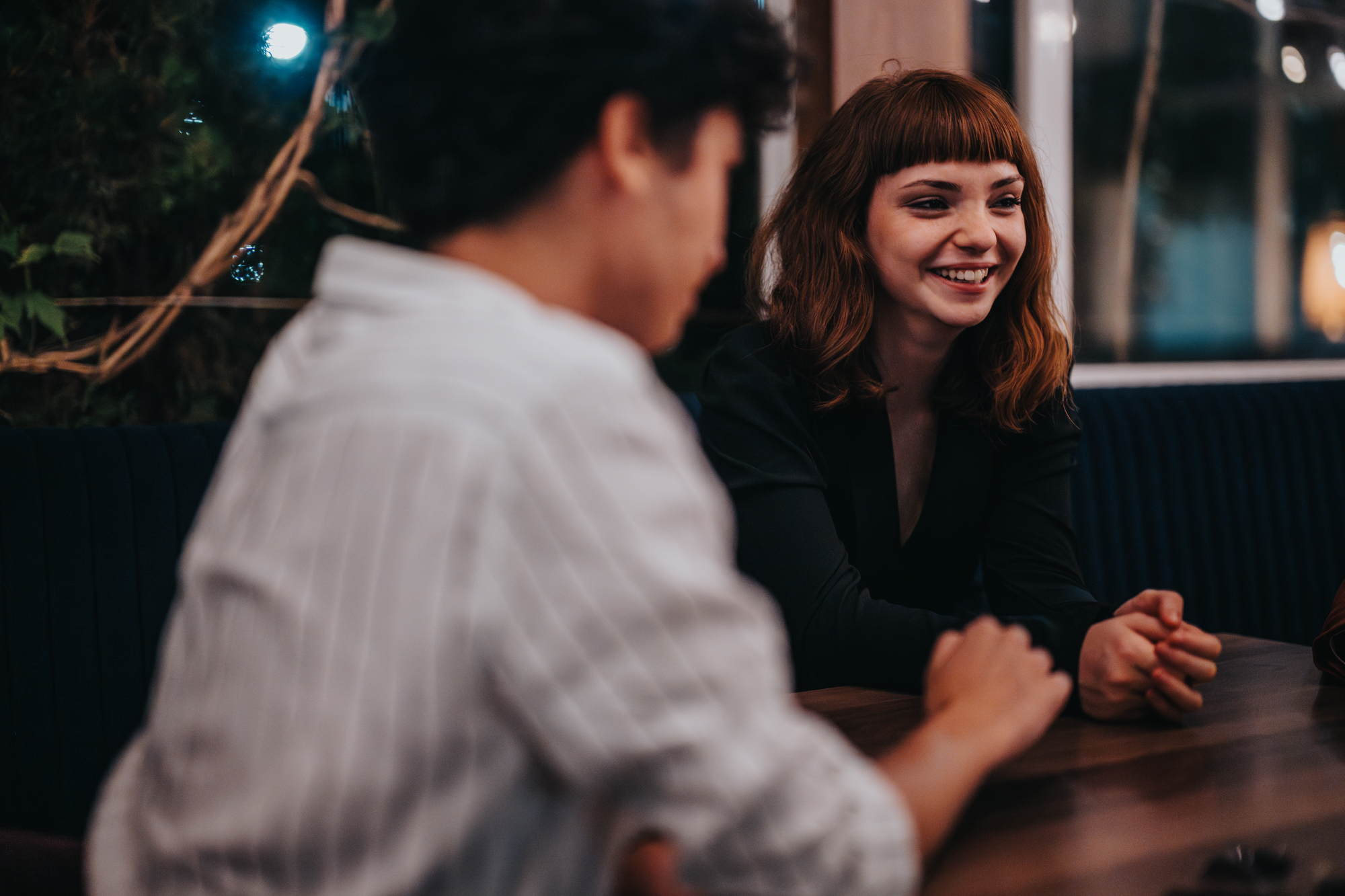 A woman with brown hair smiles while sitting at a table in a dimly lit restaurant or cafe, engaged in conversation with another person.