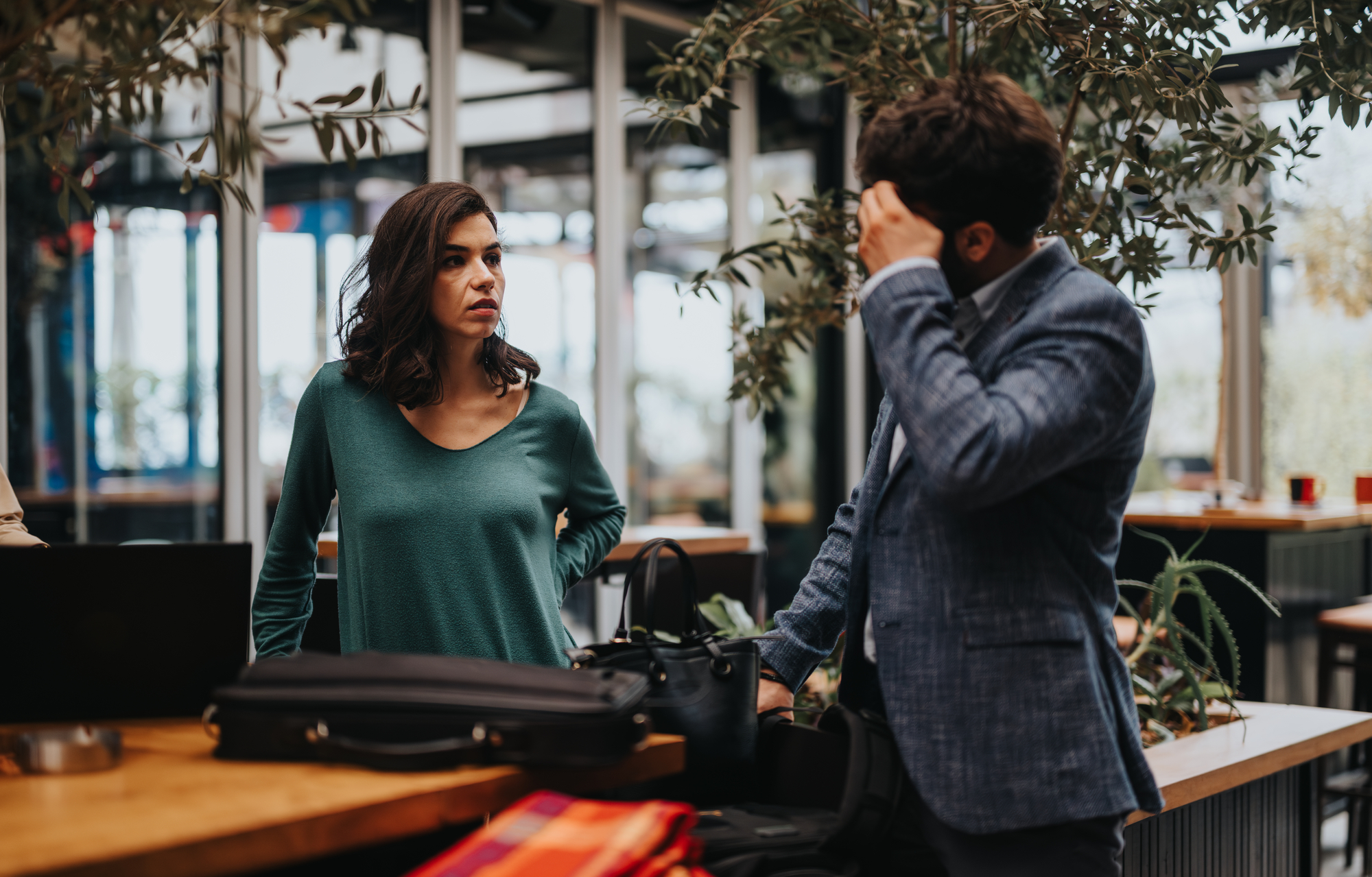 A woman in a green shirt stands with crossed arms, looking at a man in a blue suit who has his hand on his forehead. They are indoors with luggage and leafy plants around them, suggesting a travel setting.