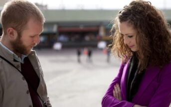 A man and woman stand outdoors in contemplation. The man with a short beard looks down, wearing a jacket and holding a camera. The woman with curly hair looks down with arms crossed, wearing a bright purple coat. They are in a blurred urban setting.