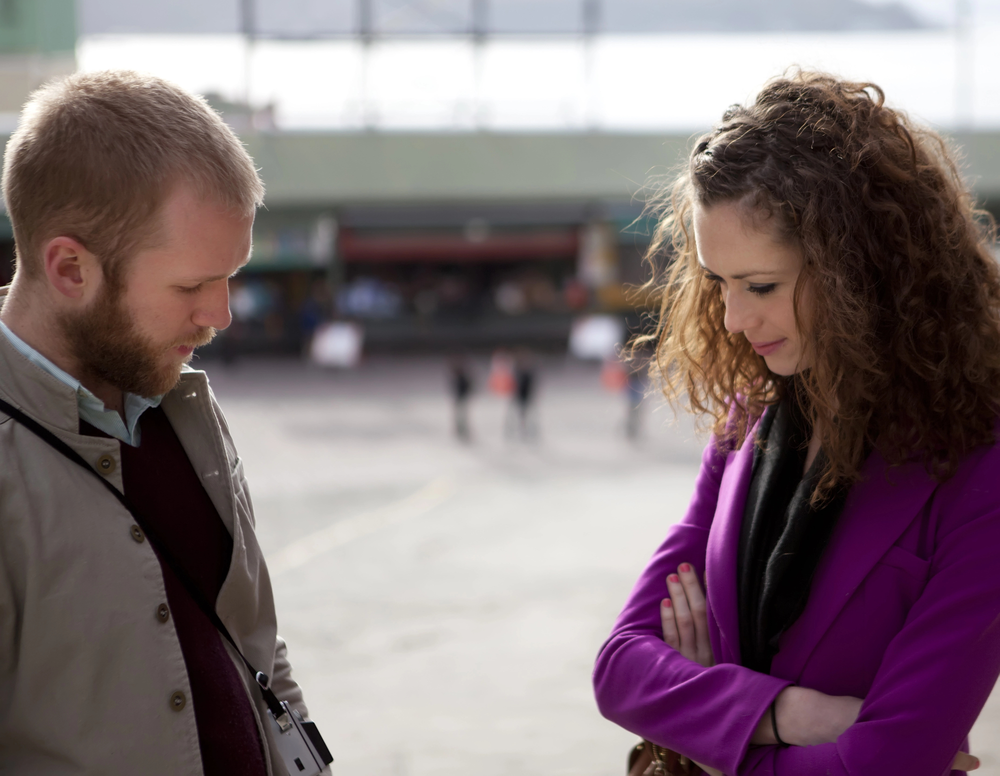 A man and woman stand outdoors in contemplation. The man with a short beard looks down, wearing a jacket and holding a camera. The woman with curly hair looks down with arms crossed, wearing a bright purple coat. They are in a blurred urban setting.