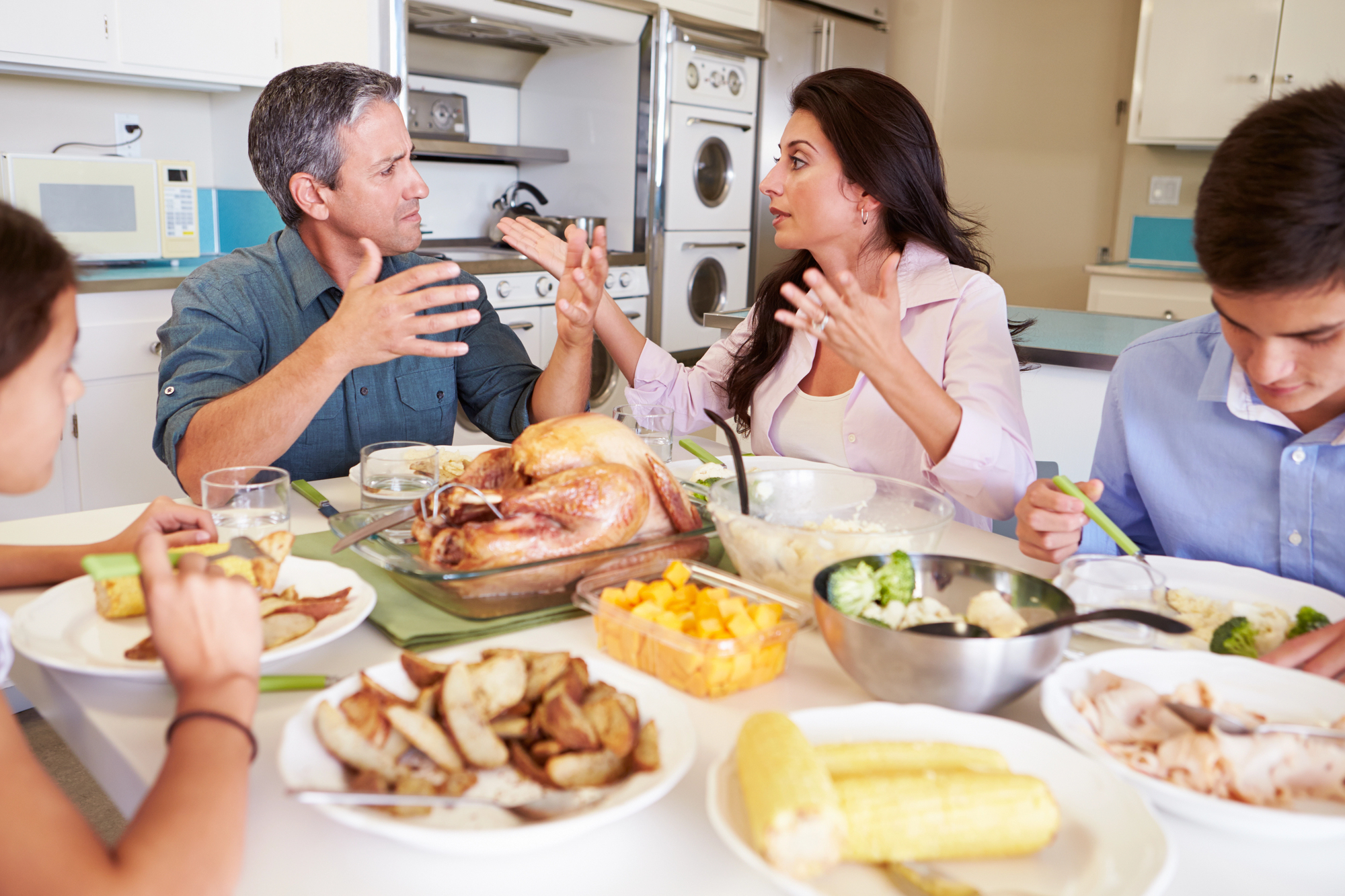 A family of four sits at a dinner table, engaged in a lively conversation. The table is set with a roasted turkey, various side dishes, and beverages. The kitchen in the background features a stove, microwave, and neatly stacked appliances.