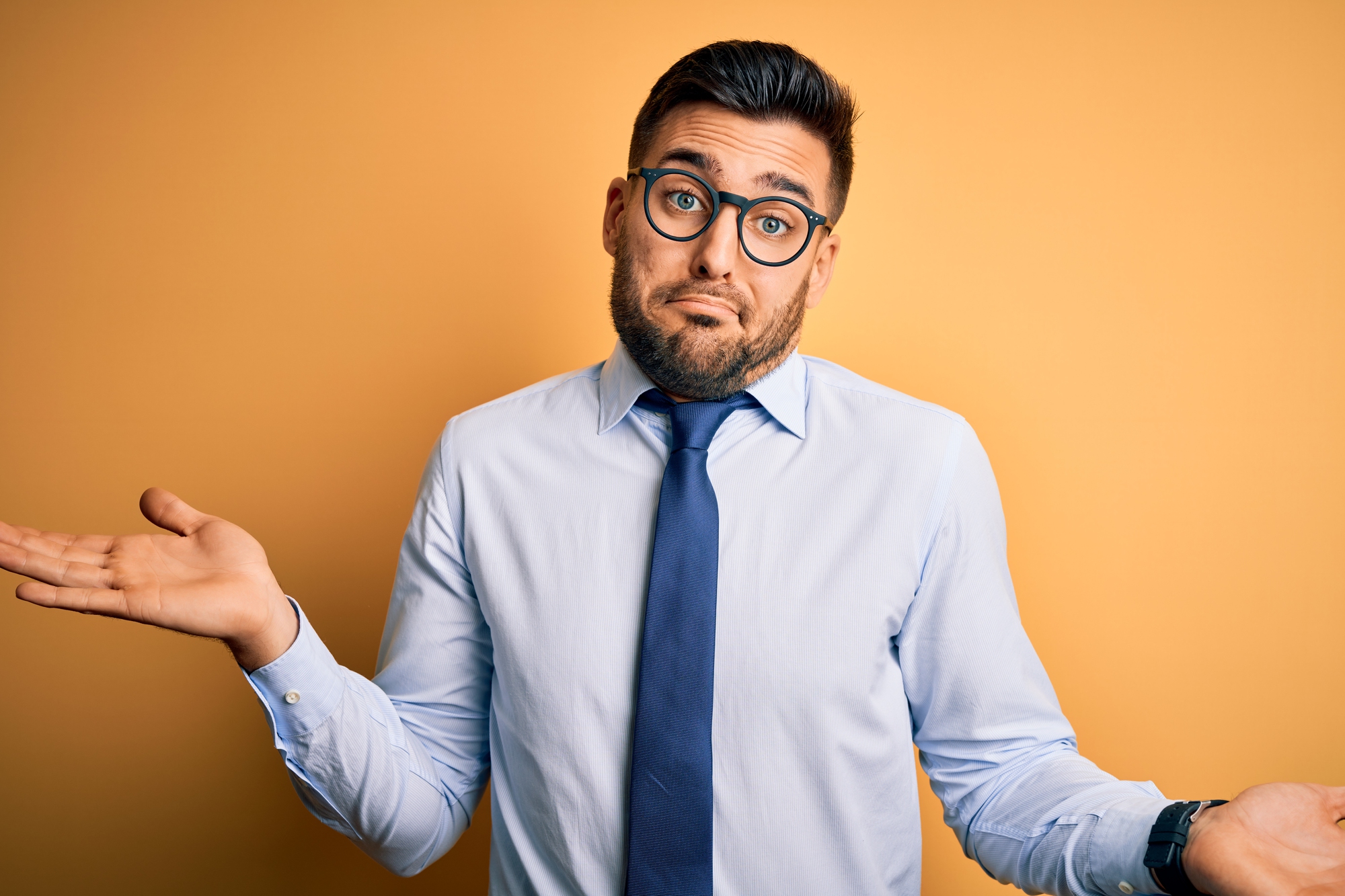 A man with a beard, wearing glasses, a light blue shirt, and a dark blue tie, stands against a mustard-colored background. He is shrugging with palms up and a puzzled expression on his face.