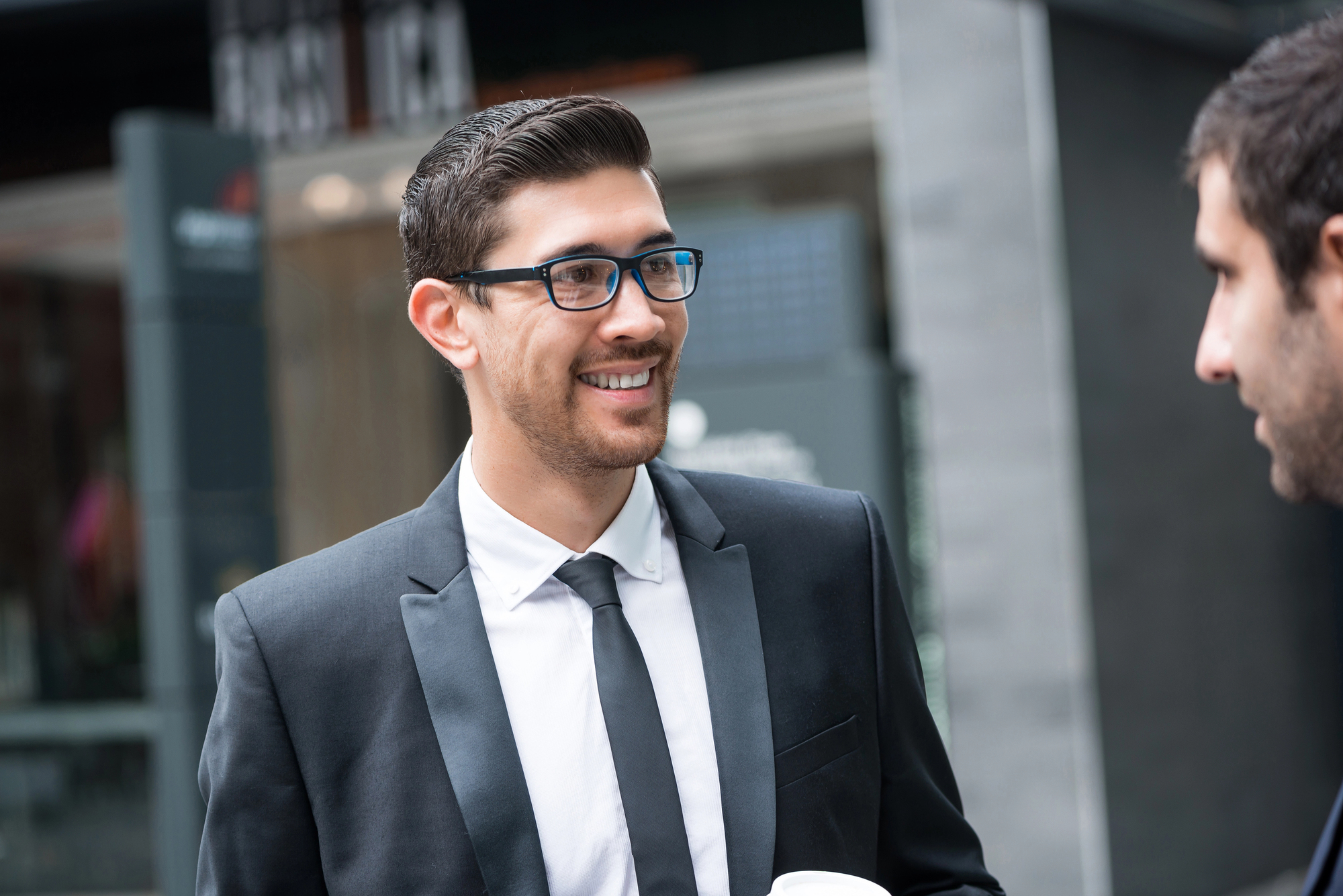 A man in a suit and tie, wearing glasses, is smiling while holding a coffee cup outdoors. He is engaged in conversation with another person out of focus in the foreground. The background shows an urban setting with a modern building.