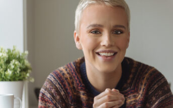 A person with short blonde hair smiles warmly at the camera, wearing a patterned sweater. They are sitting indoors with a small potted plant and a white mug in the background.