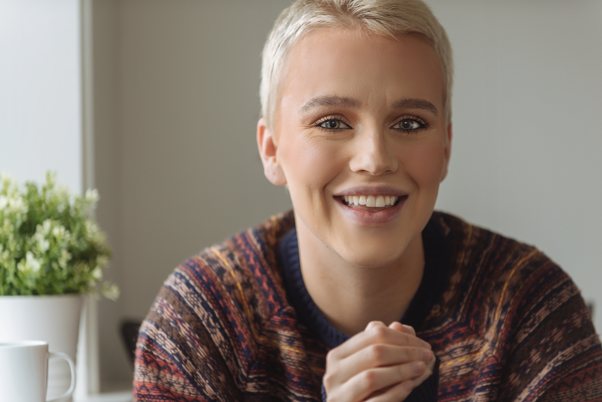 A person with short blonde hair smiles warmly at the camera, wearing a patterned sweater. They are sitting indoors with a small potted plant and a white mug in the background.