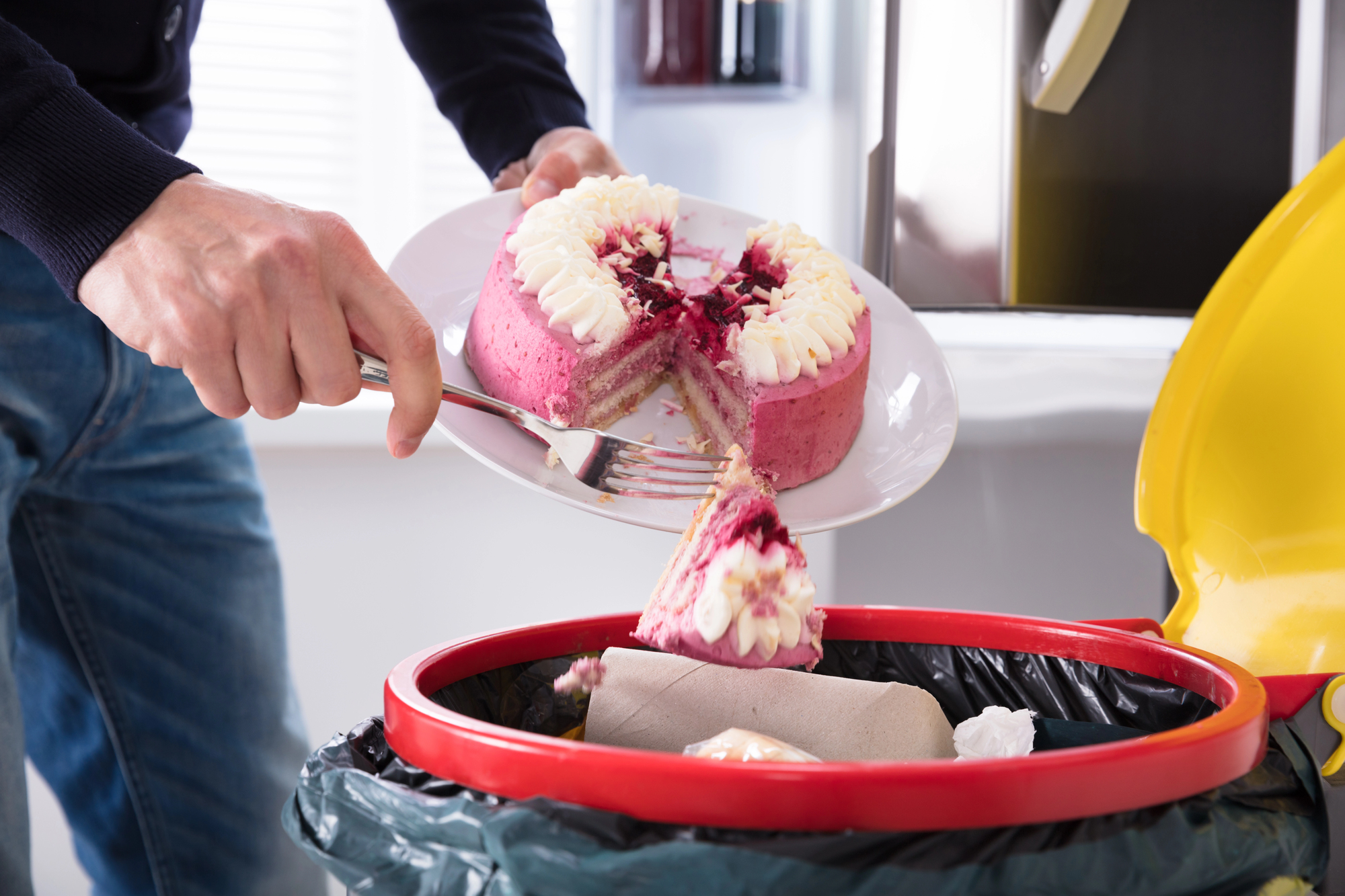 A person discards a partially eaten pink cake with white frosting into a red trash bin. The cake is on a white plate, and the person is using a fork to remove a slice. The scene is indoors, with part of a kitchen visible in the background.