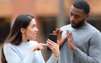 A woman and a man are outdoors. The woman, holding a smartphone, looks upset as she talks to the man, who appears surprised and raises his hands defensively. Both are wearing casual sweaters.