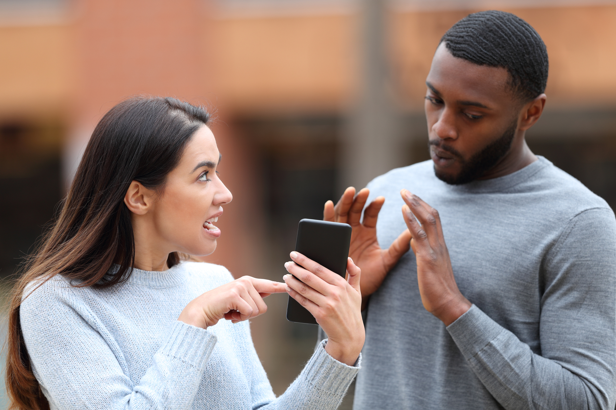 A woman and a man are outdoors. The woman, holding a smartphone, looks upset as she talks to the man, who appears surprised and raises his hands defensively. Both are wearing casual sweaters.