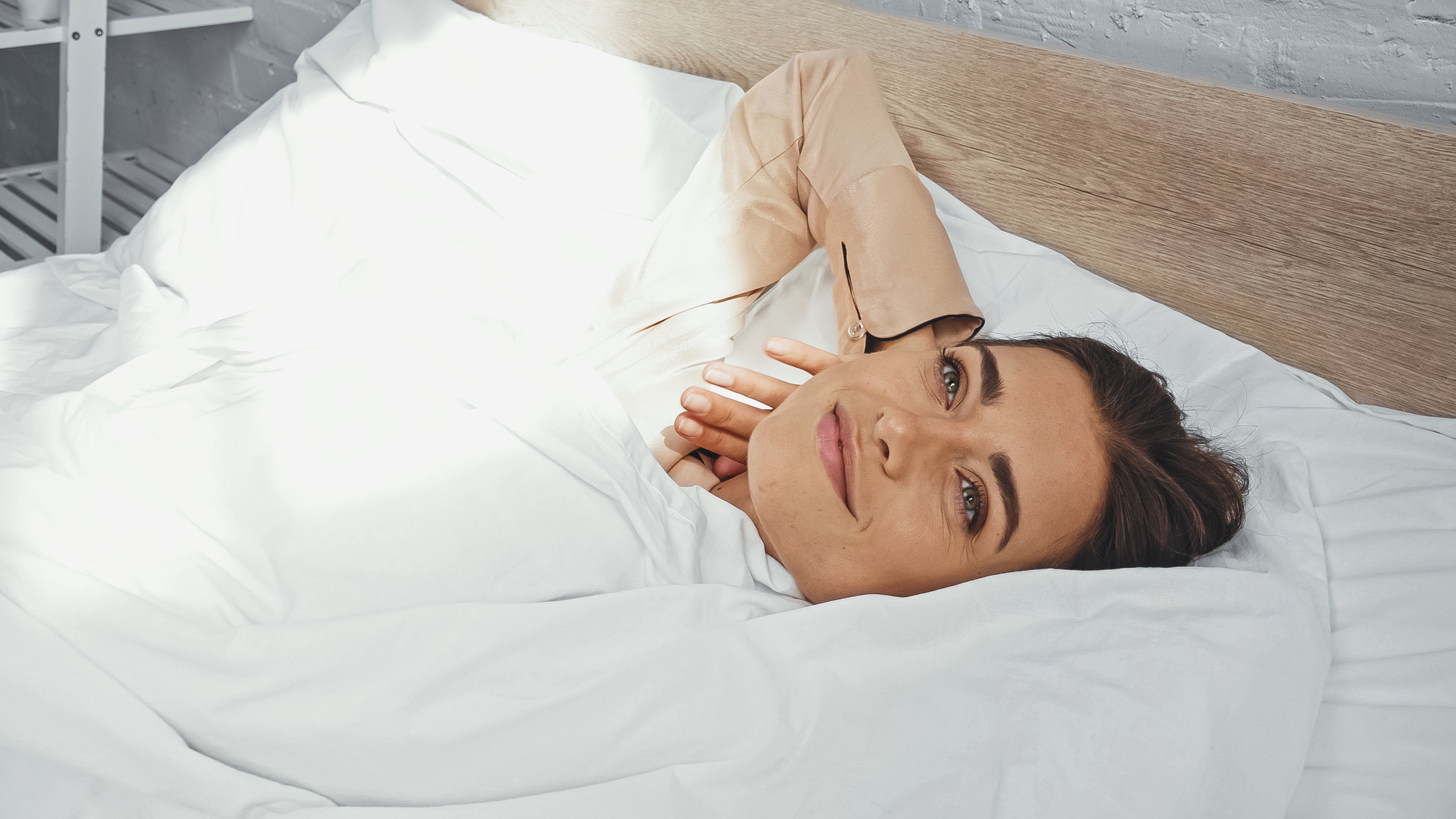 A person with dark hair lying in bed, partially covered by a white blanket. They are resting their head on one hand and smiling softly, with sunlight streaming onto the bedding. The headboard is wooden, and the room has a calm atmosphere.