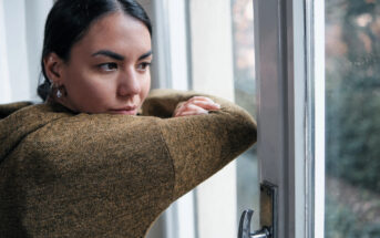 A person in a brown sweater leans on a windowsill, gazing outside with a thoughtful expression. The window is slightly open, and soft natural light illuminates their face. Trees and greenery are visible in the background.