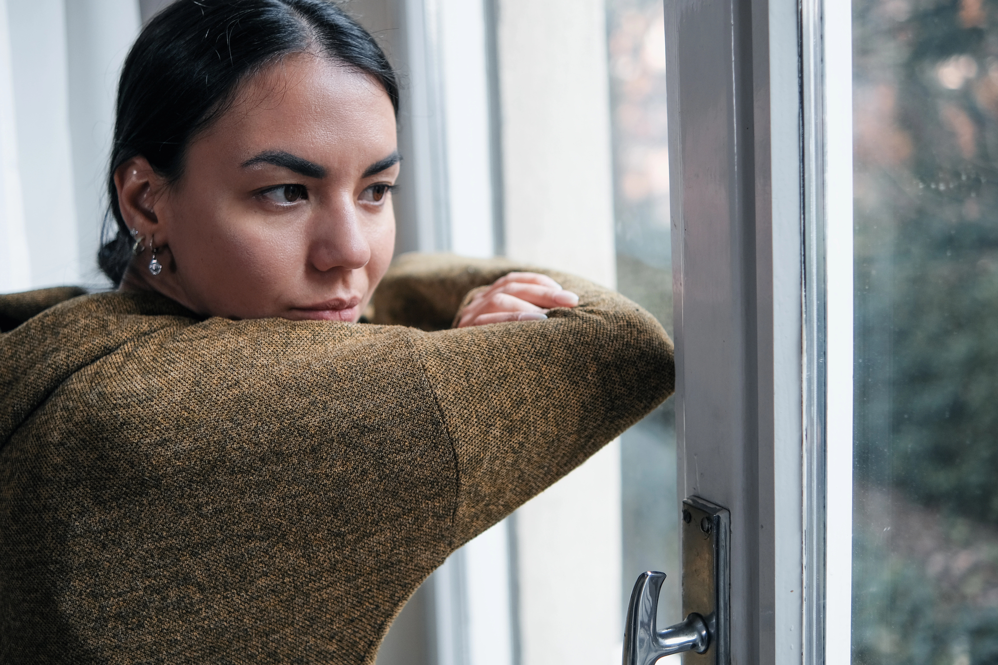 A person in a brown sweater leans on a windowsill, gazing outside with a thoughtful expression. The window is slightly open, and soft natural light illuminates their face. Trees and greenery are visible in the background.