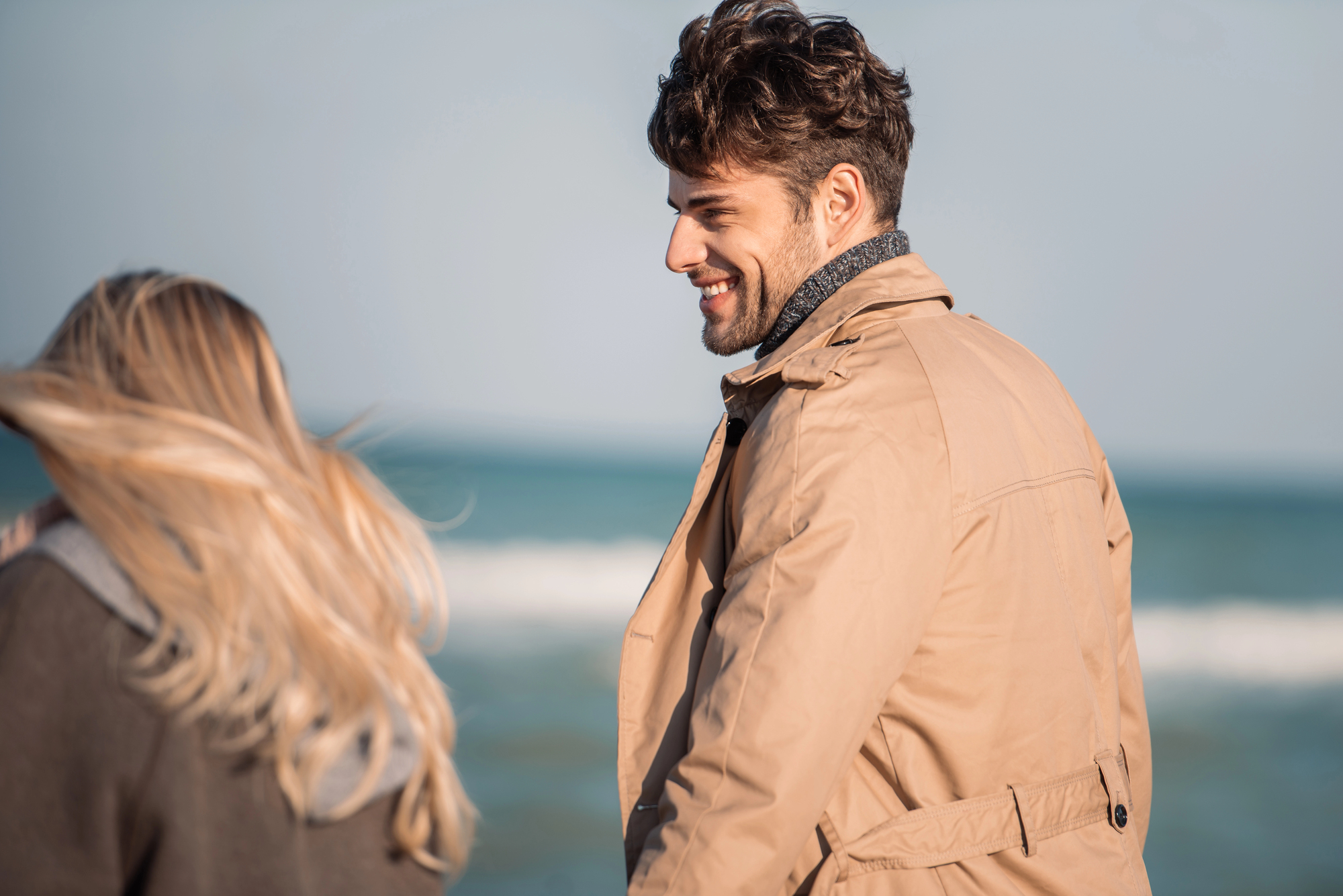 A man in a tan coat smiles while standing by the sea. A woman with long blonde hair faces away from the camera, both enjoying a breezy day at the beach.