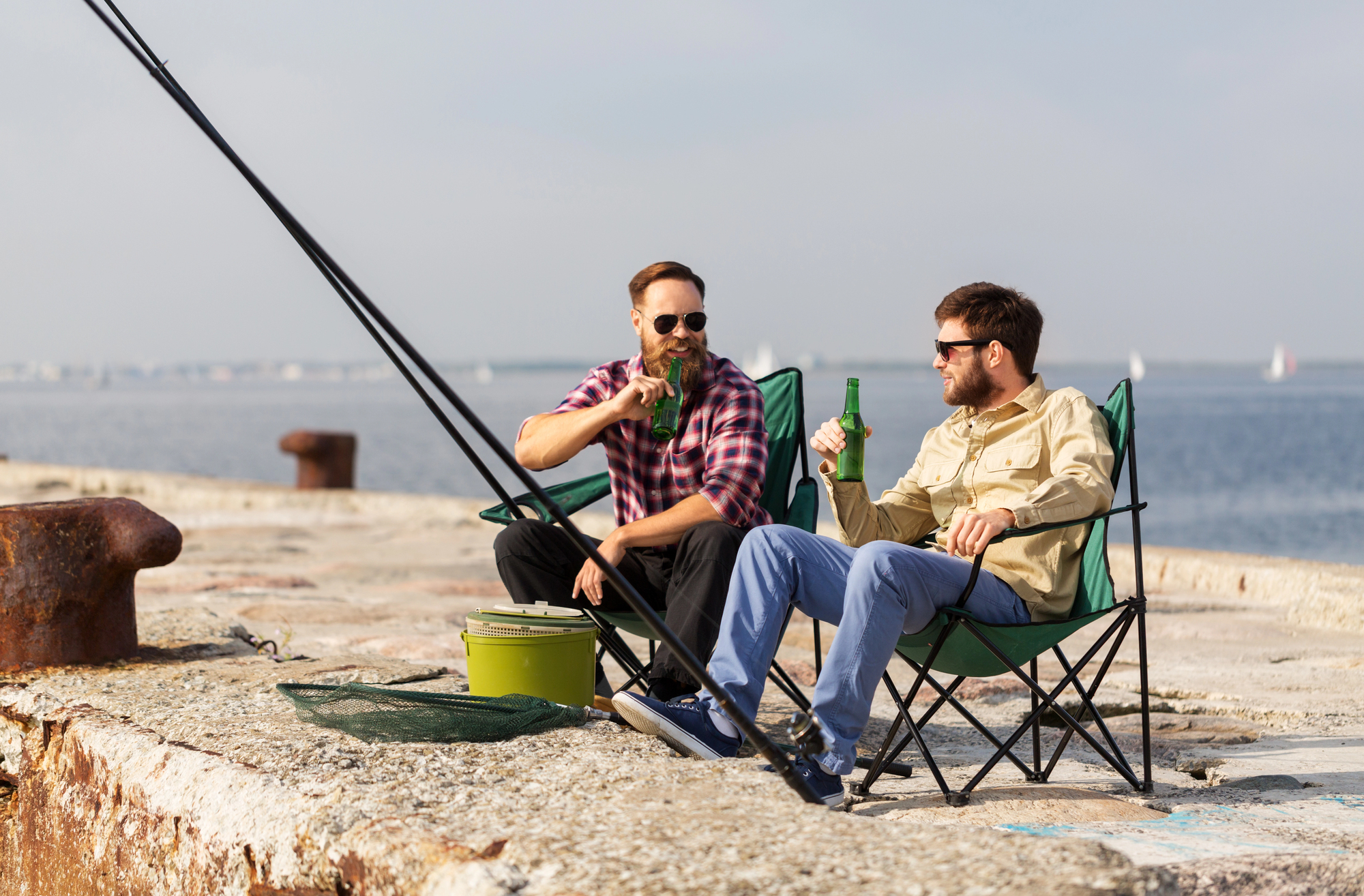 Two men sitting on folding chairs by the seaside, enjoying beverages. One is wearing sunglasses and holding a fishing rod. A bucket and net are beside them. The ocean and sailboats are visible in the background under a clear sky.