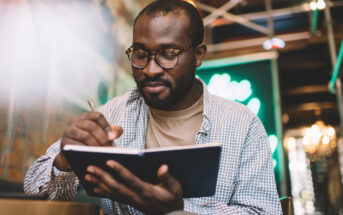 A person with glasses and a checkered shirt is sitting indoors, writing in a notebook. The background is blurred with some green and yellow lights. The scene appears calm and focused.