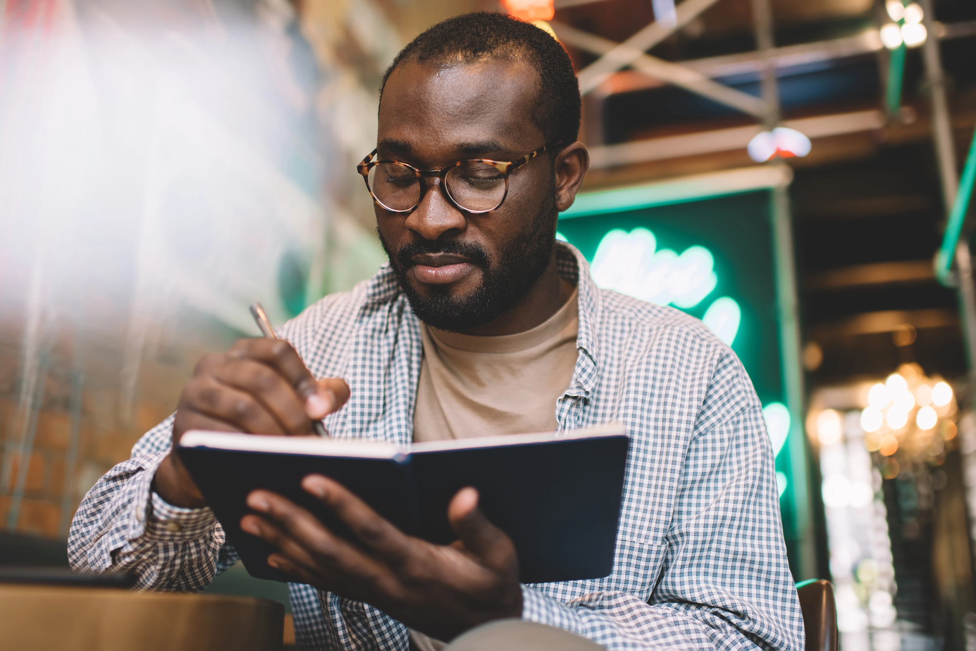 A person with glasses and a checkered shirt is sitting indoors, writing in a notebook. The background is blurred with some green and yellow lights. The scene appears calm and focused.