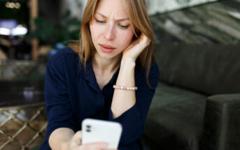 A woman with long hair looks at her smartphone with a concerned expression, sitting on a sofa in a modern room. She is wearing a navy blouse and a beaded bracelet, resting her chin on her hand.