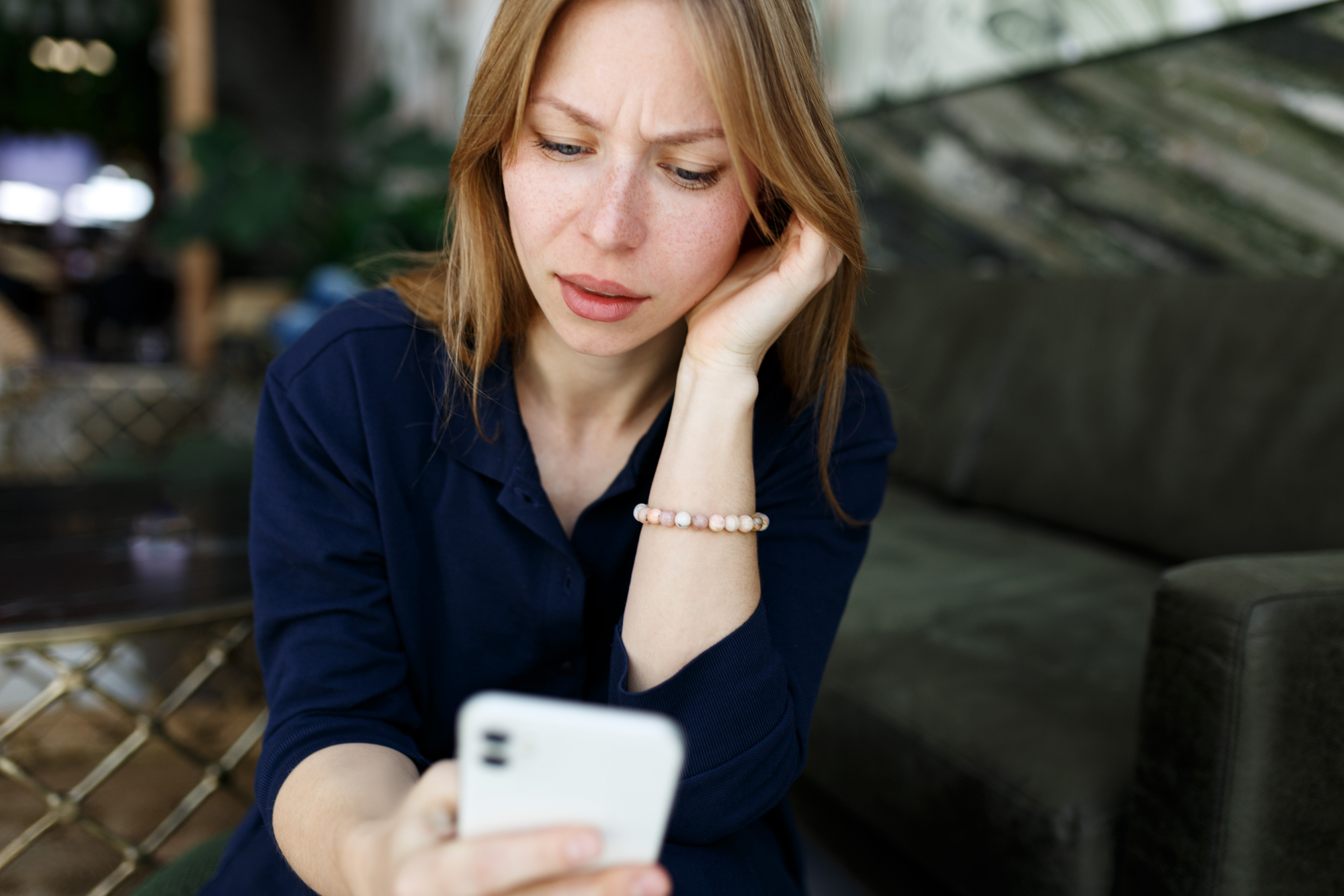 A woman with long hair looks at her smartphone with a concerned expression, sitting on a sofa in a modern room. She is wearing a navy blouse and a beaded bracelet, resting her chin on her hand.