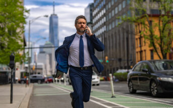 A man in a blue suit is running down a city street while talking on his phone. He appears focused, with buildings and a busy road in the background. It's a sunny day, and there are cars and trees along the street.