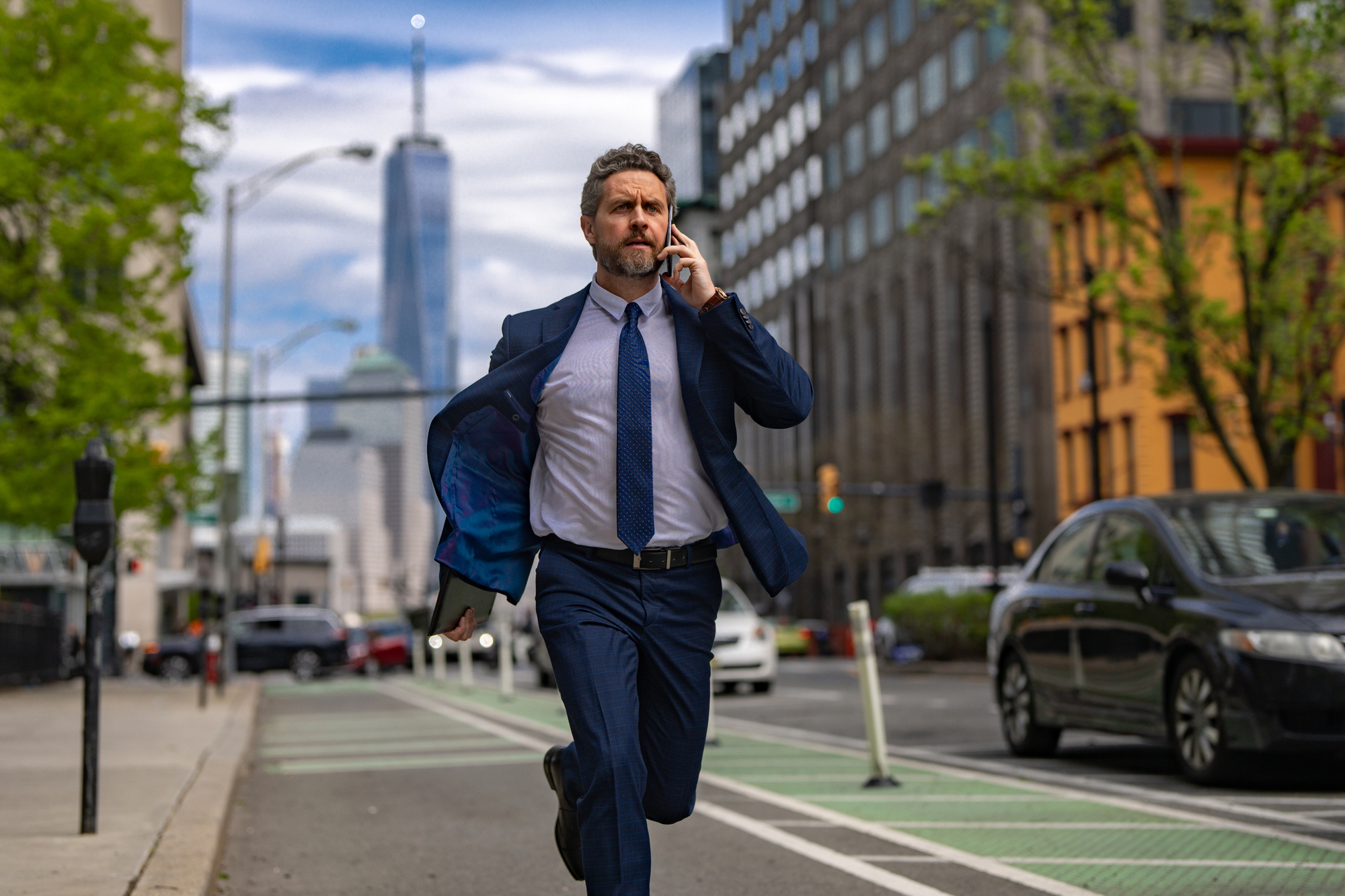 A man in a blue suit is running down a city street while talking on his phone. He appears focused, with buildings and a busy road in the background. It's a sunny day, and there are cars and trees along the street.