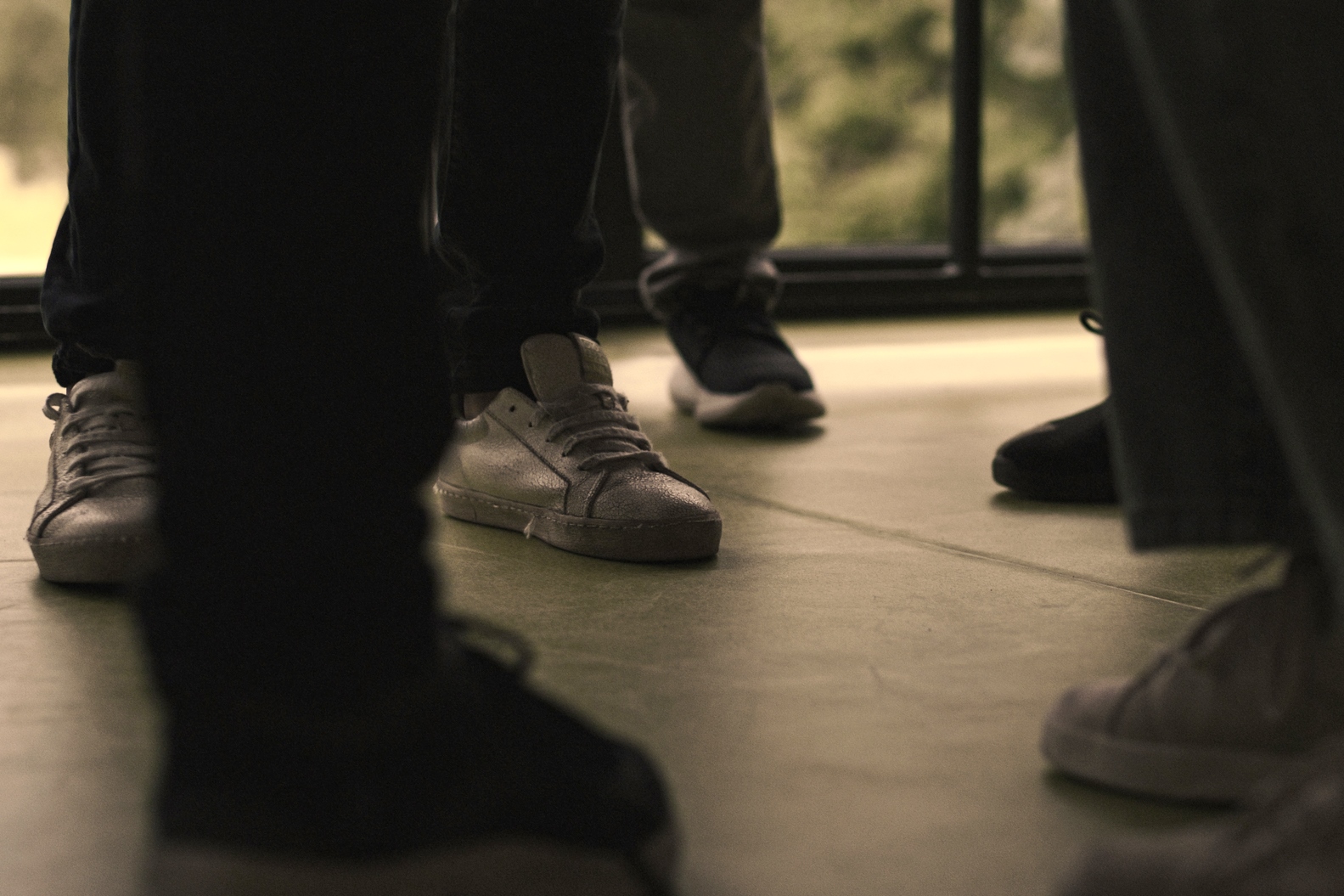 Several people stand together indoors, viewed from the knees down. They are wearing casual sneakers, and the floor appears to be smooth and light-colored. The background shows a window with a blurred view outside.