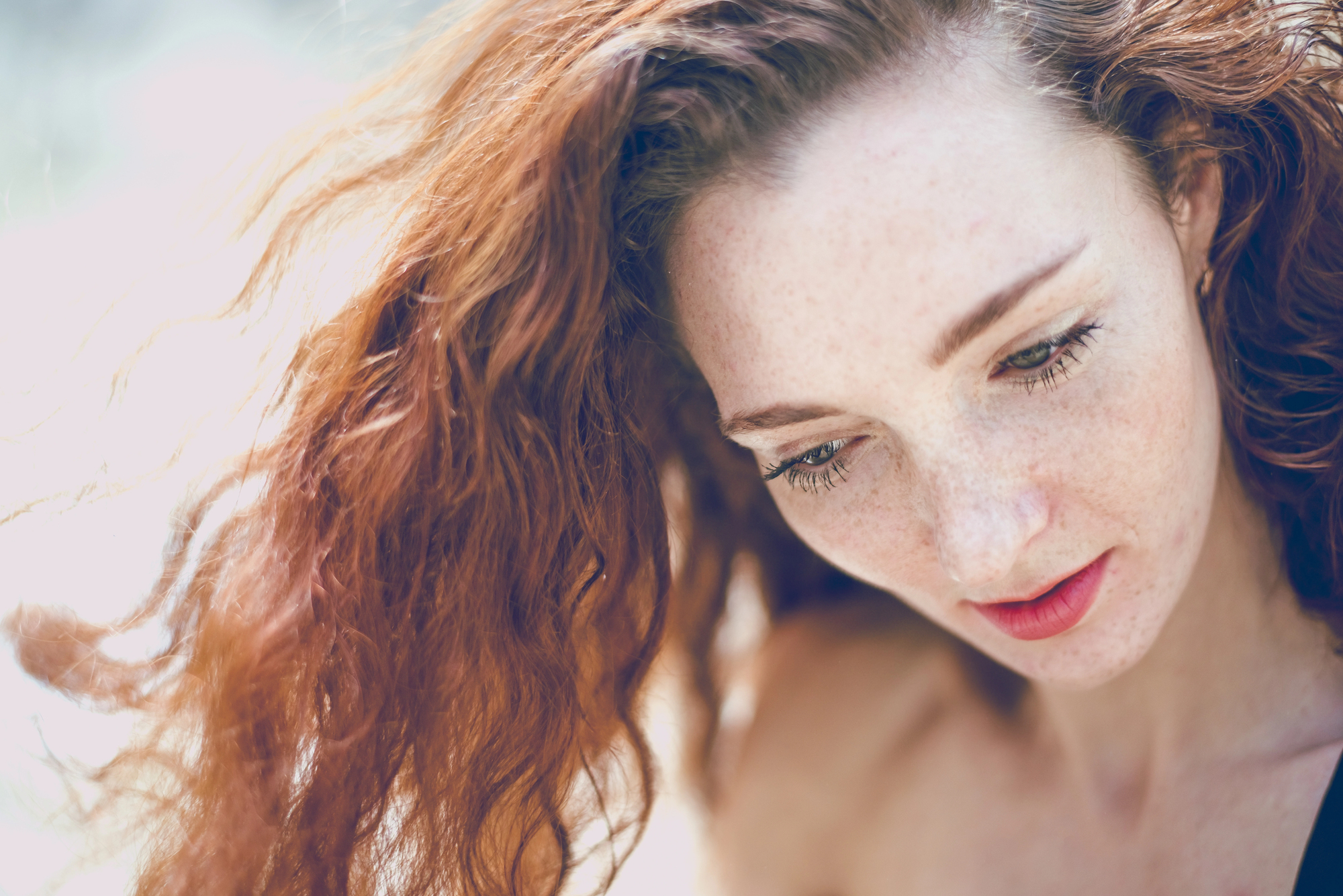 A woman with long, wavy red hair and freckles looks down. She has fair skin and is wearing light makeup, with red lipstick. The background is softly blurred, highlighting her contemplative expression.