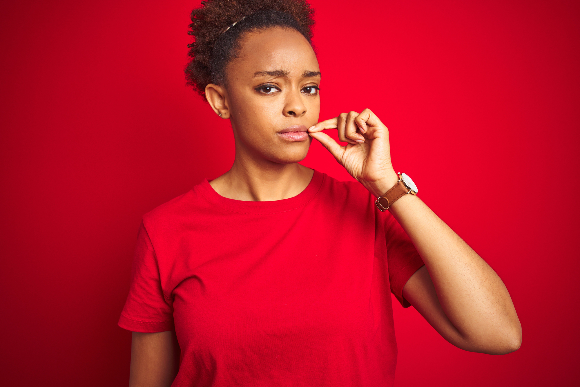 A person wearing a red shirt stands against a red background, looking thoughtful while biting their lip and holding a finger to it. They have short curly hair and are wearing a watch on their wrist.