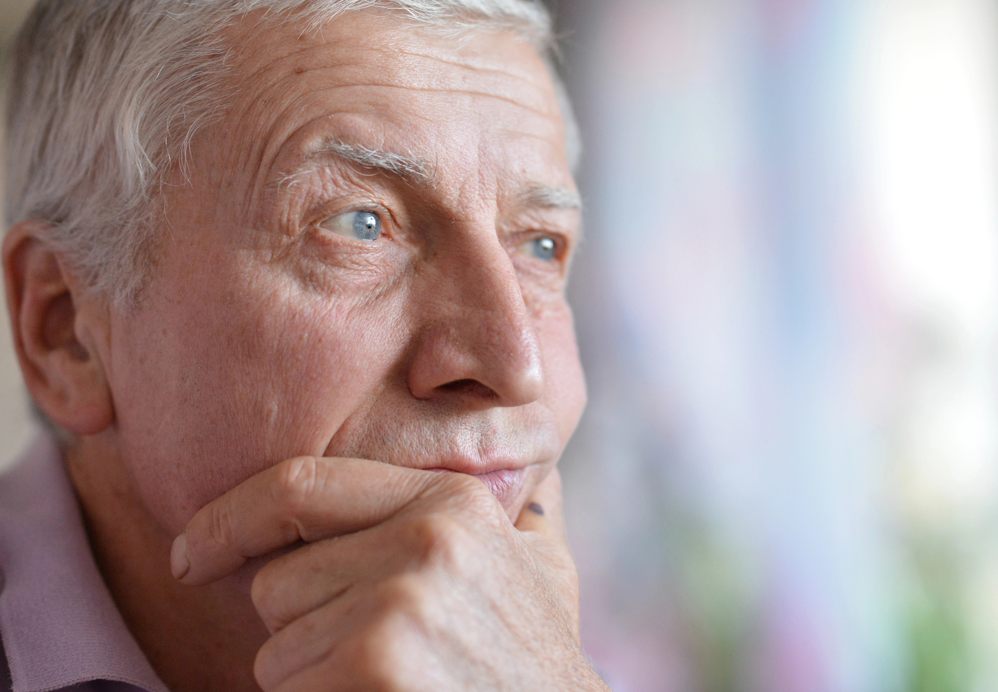 A close-up of an elderly man with gray hair, looking thoughtfully into the distance. He rests his chin on his hand, with a soft expression on his face. The background is blurred, suggesting an indoor setting.