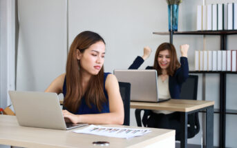 Two women are in an office, each sitting at a desk with a laptop. The woman in the foreground looks frustrated and is gazing at her screen. In the background, the woman appears excited and is raising her fists in triumph. Shelves with books are visible.