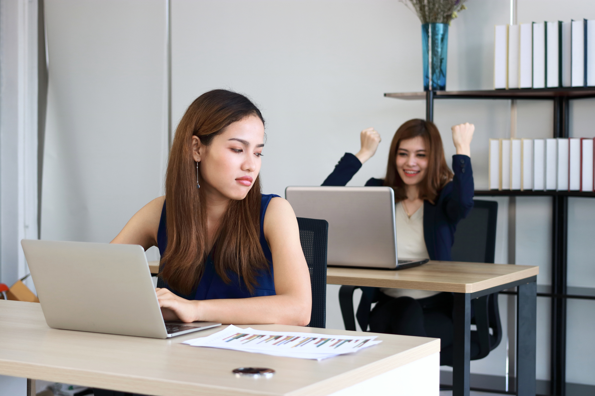 Two women are in an office, each sitting at a desk with a laptop. The woman in the foreground looks frustrated and is gazing at her screen. In the background, the woman appears excited and is raising her fists in triumph. Shelves with books are visible.