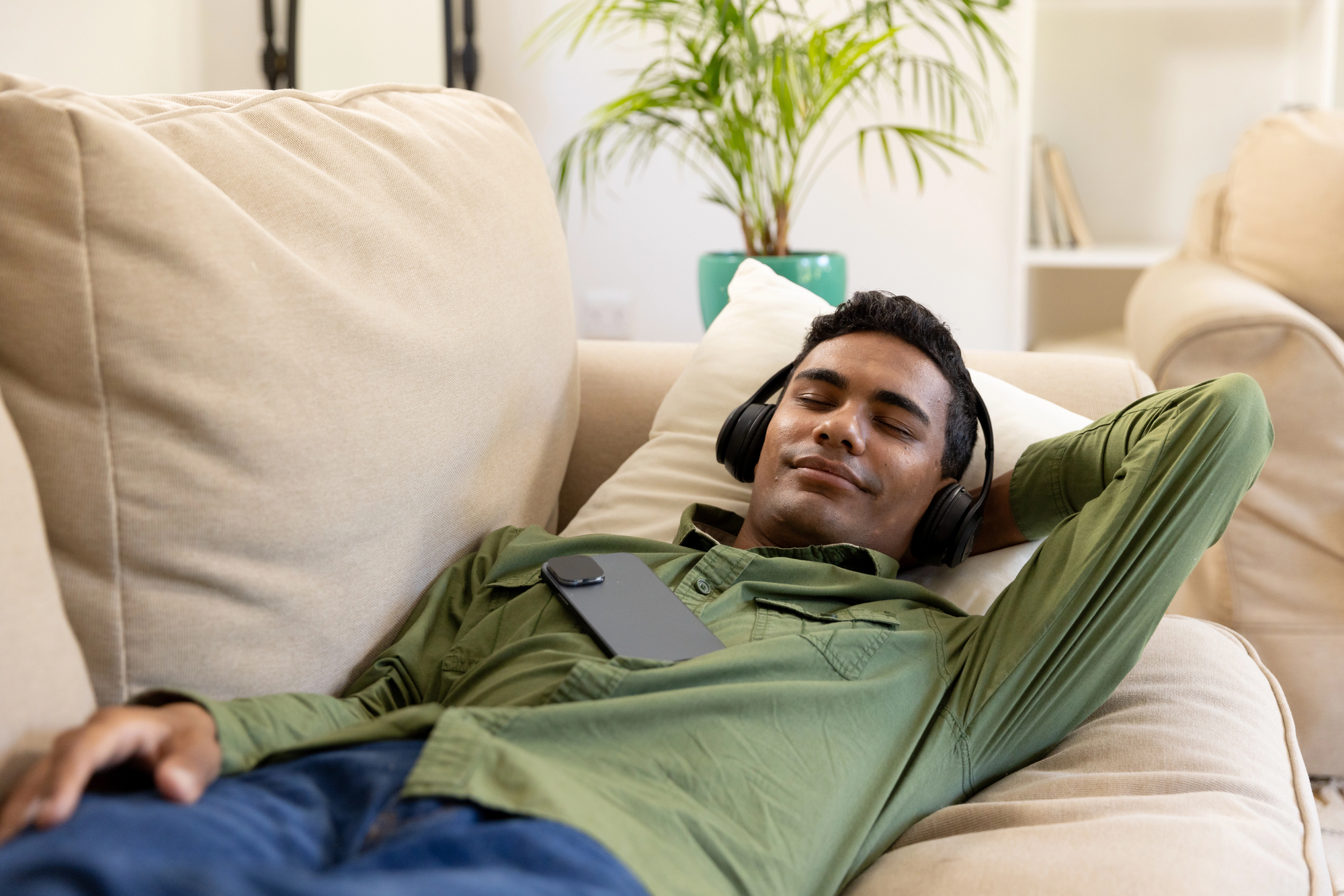 A person is relaxing on a beige couch, wearing headphones and a green shirt. They have a smartphone resting on their chest and appear to be enjoying music. In the background, there is a potted plant and a bookshelf.