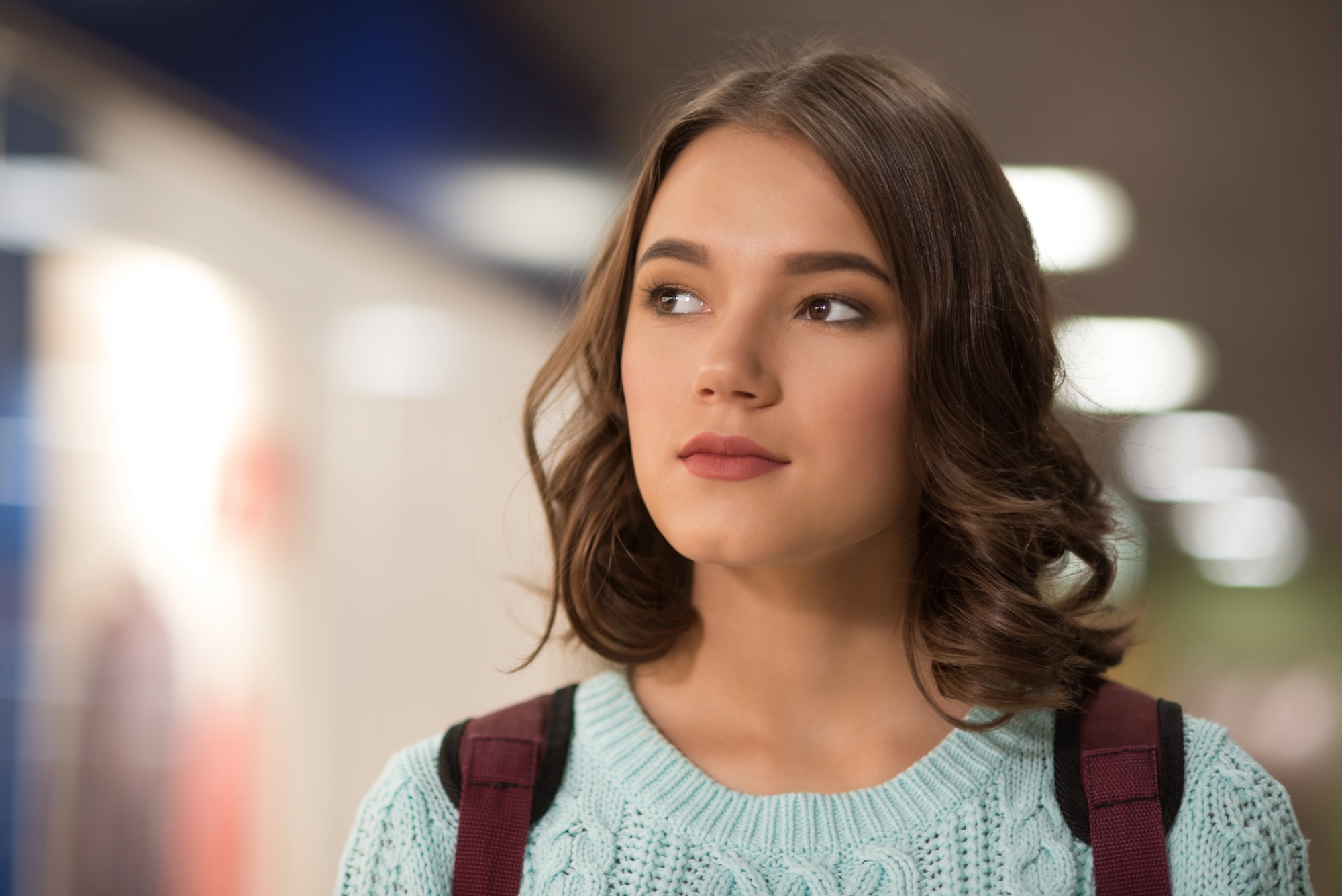 A young woman with brown hair and a thoughtful expression stands indoors. She is wearing a light blue sweater and has a maroon strap over her shoulders. The background is softly blurred with lights and hints of color.