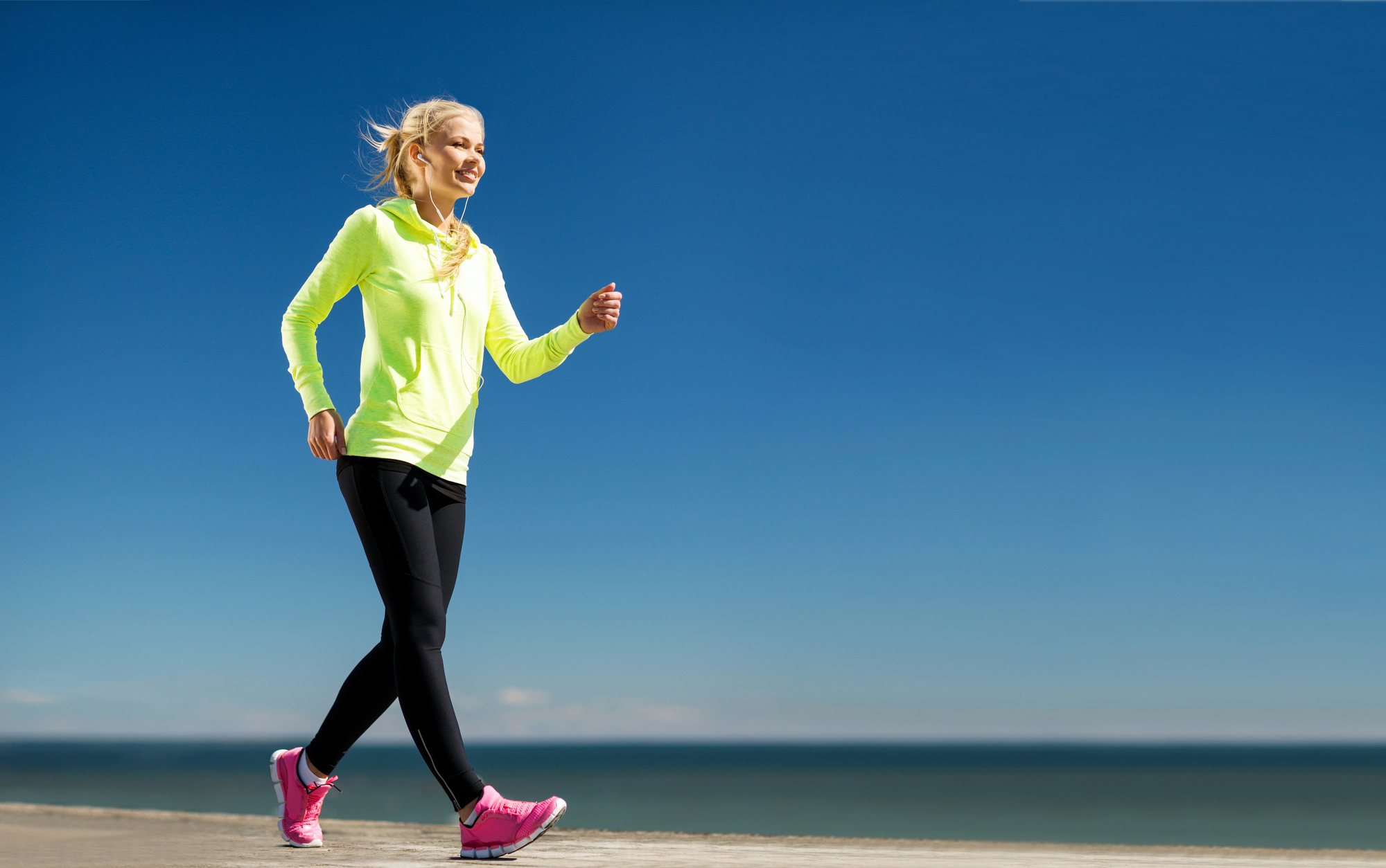 A person in a bright yellow hoodie and black leggings is walking along a beachside pathway. The sky is clear and blue, and the ocean is visible in the background. They are wearing pink sneakers.