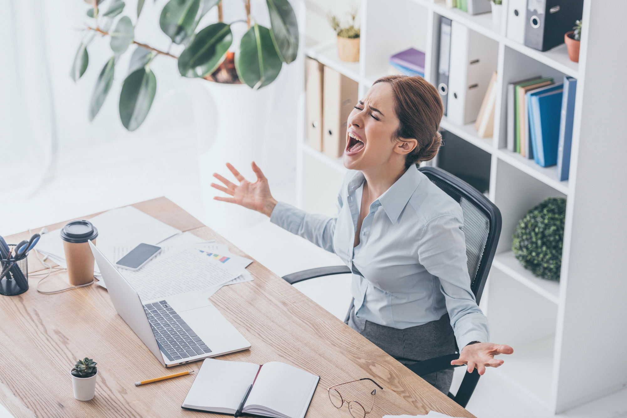 A woman at a desk with a laptop and office supplies appears frustrated, raising her arms and shouting. She is sitting in a modern, well-lit office space with bookshelves and a large plant in the background.