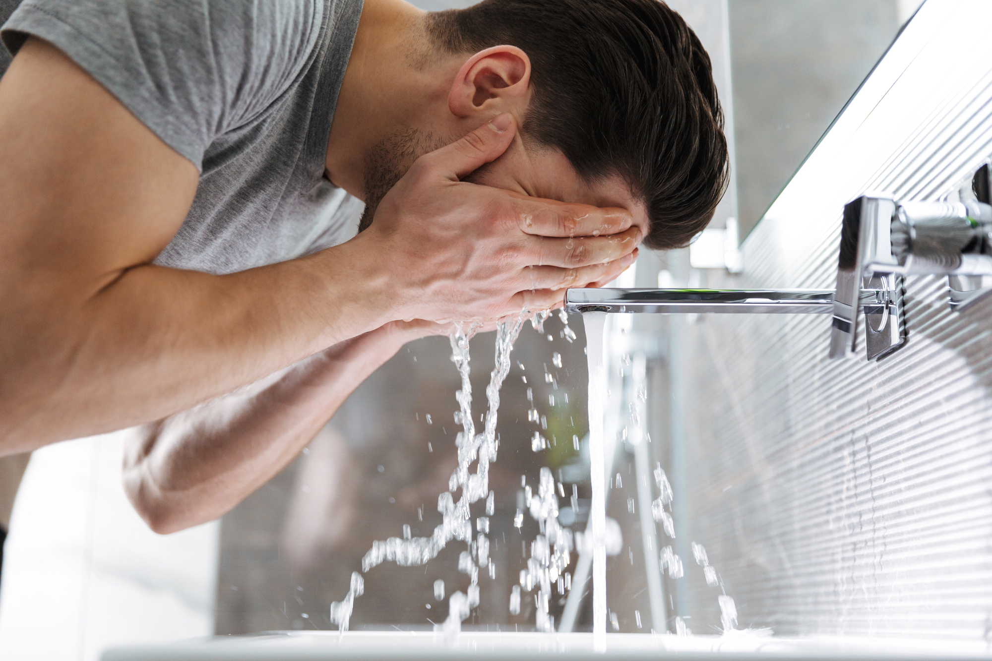 A person in a gray shirt washes their face with water from a modern faucet in a bright bathroom. The image captures water splashing as they lean over the sink.