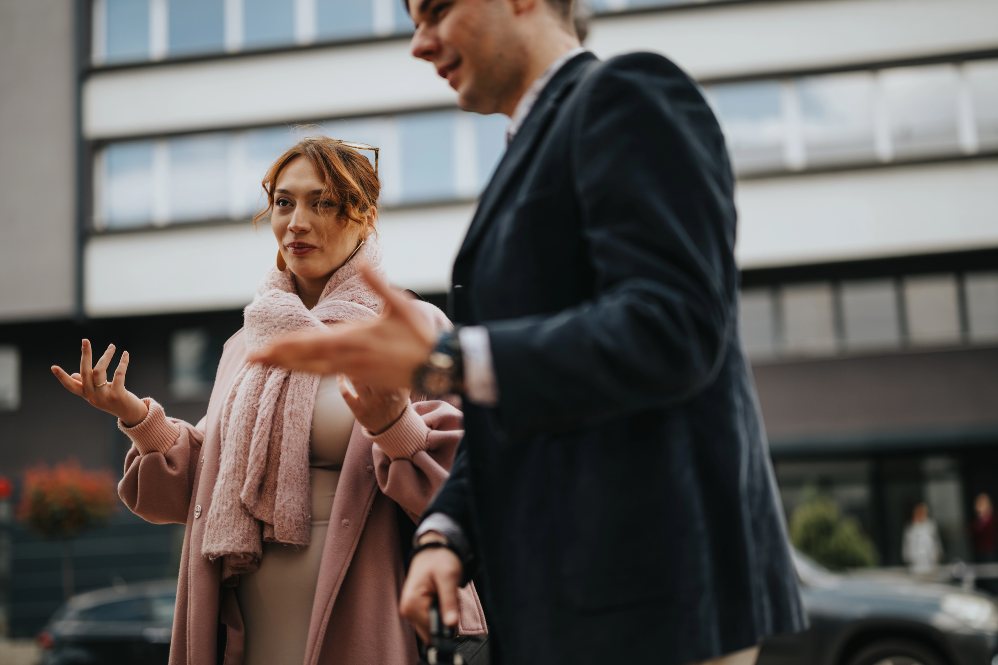 A woman and a man in conversation outdoors. The woman wears a pink coat and scarf, gesturing with her hands. The man, in a dark suit, faces her. A modern building is in the background.