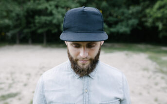 A man with a beard wearing a black cap and light-colored button-up shirt looks down while standing outdoors. The background features blurred greenery and sand.