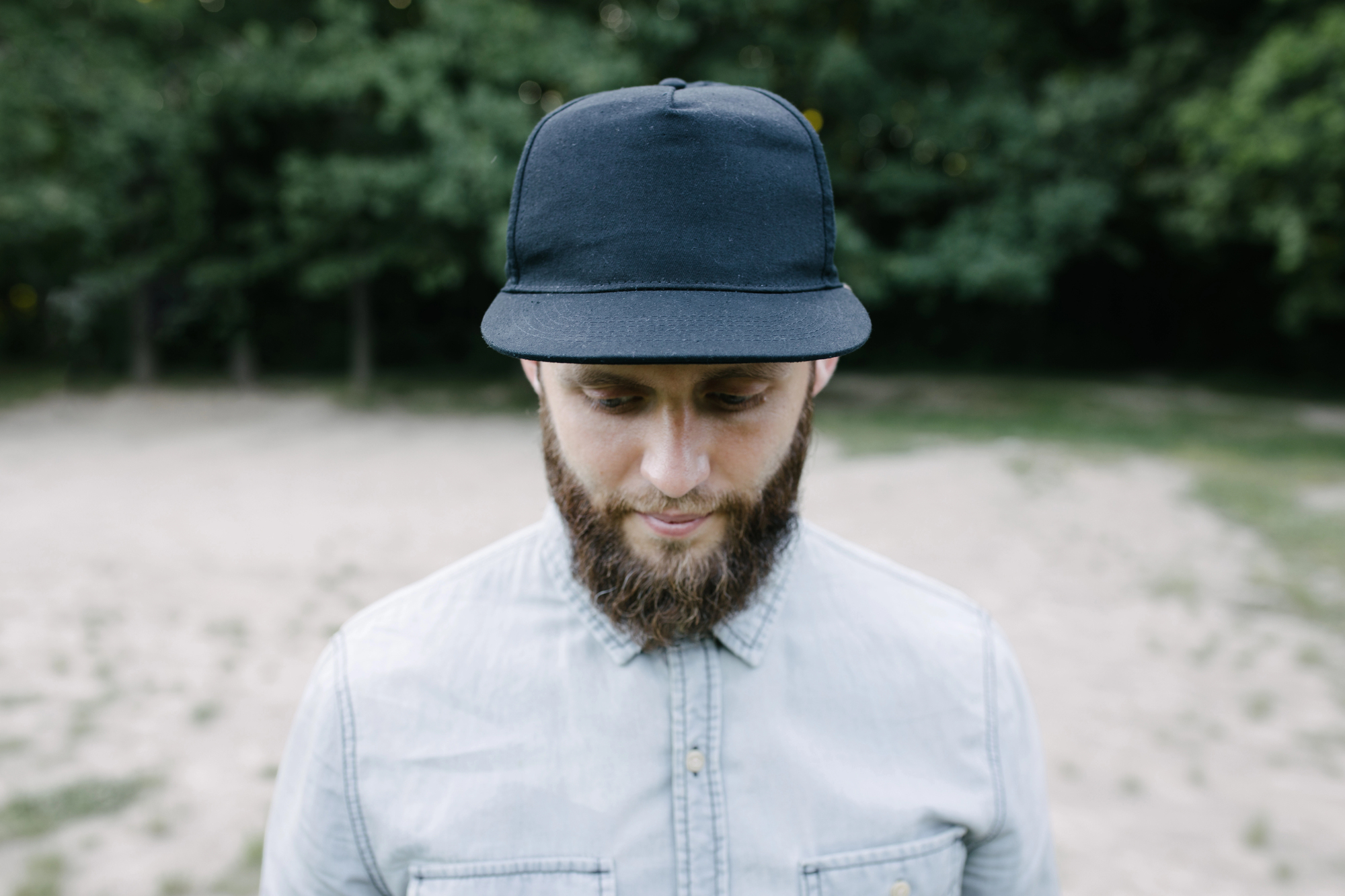 A man with a beard wearing a black cap and light-colored button-up shirt looks down while standing outdoors. The background features blurred greenery and sand.