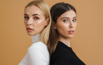 Two women standing back-to-back against a beige background. The woman on the left has long blonde hair and wears a white turtleneck. The woman on the right has long brown hair and wears a black turtleneck. Both look directly at the camera.