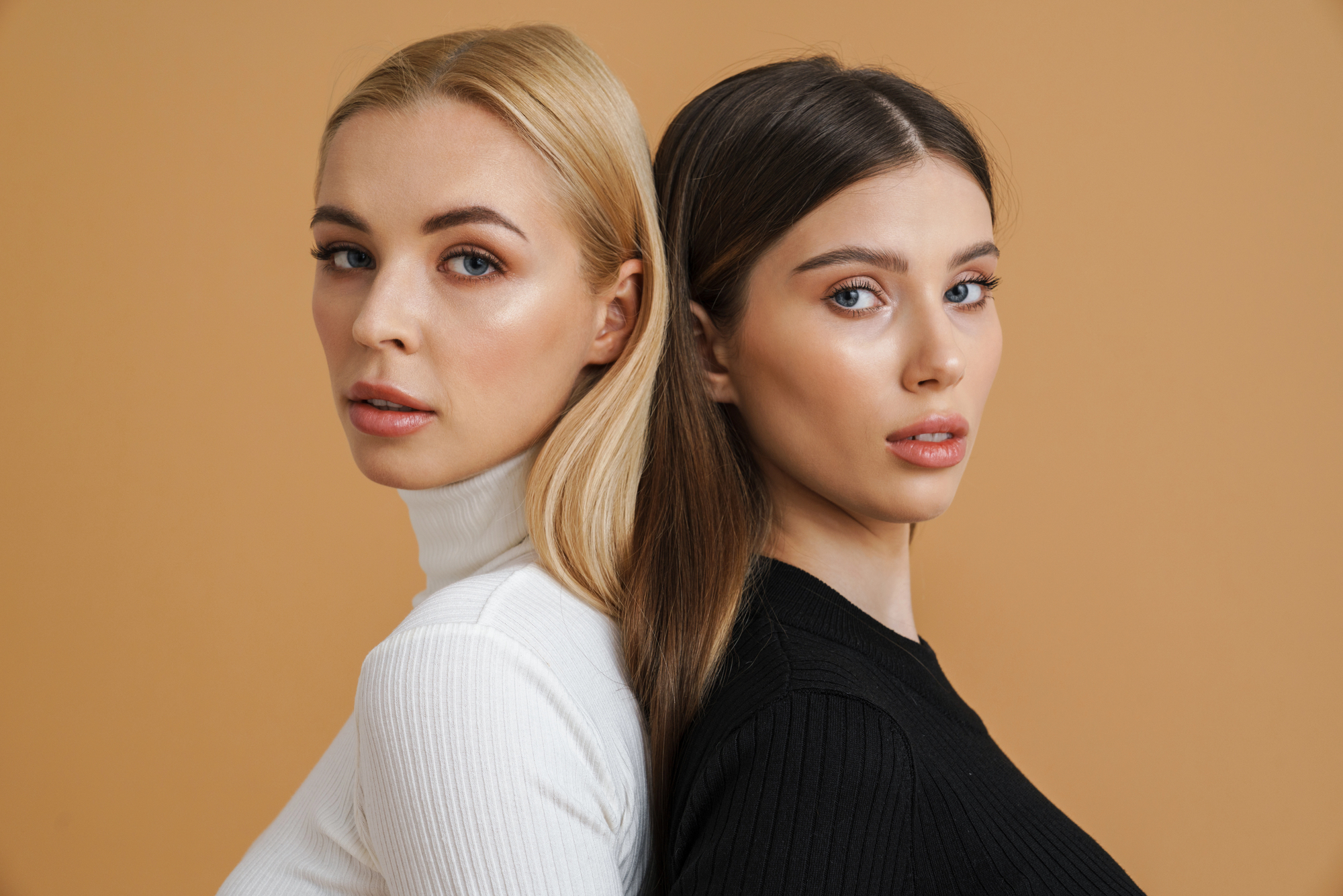 Two women standing back-to-back against a beige background. The woman on the left has long blonde hair and wears a white turtleneck. The woman on the right has long brown hair and wears a black turtleneck. Both look directly at the camera.