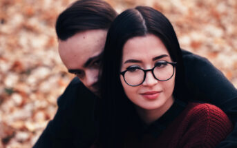 A woman with glasses and dark hair, wearing a red sweater, is sitting in front of a man with dark hair. They are outdoors with autumn leaves on the ground. The man partially hides his face behind her shoulder.