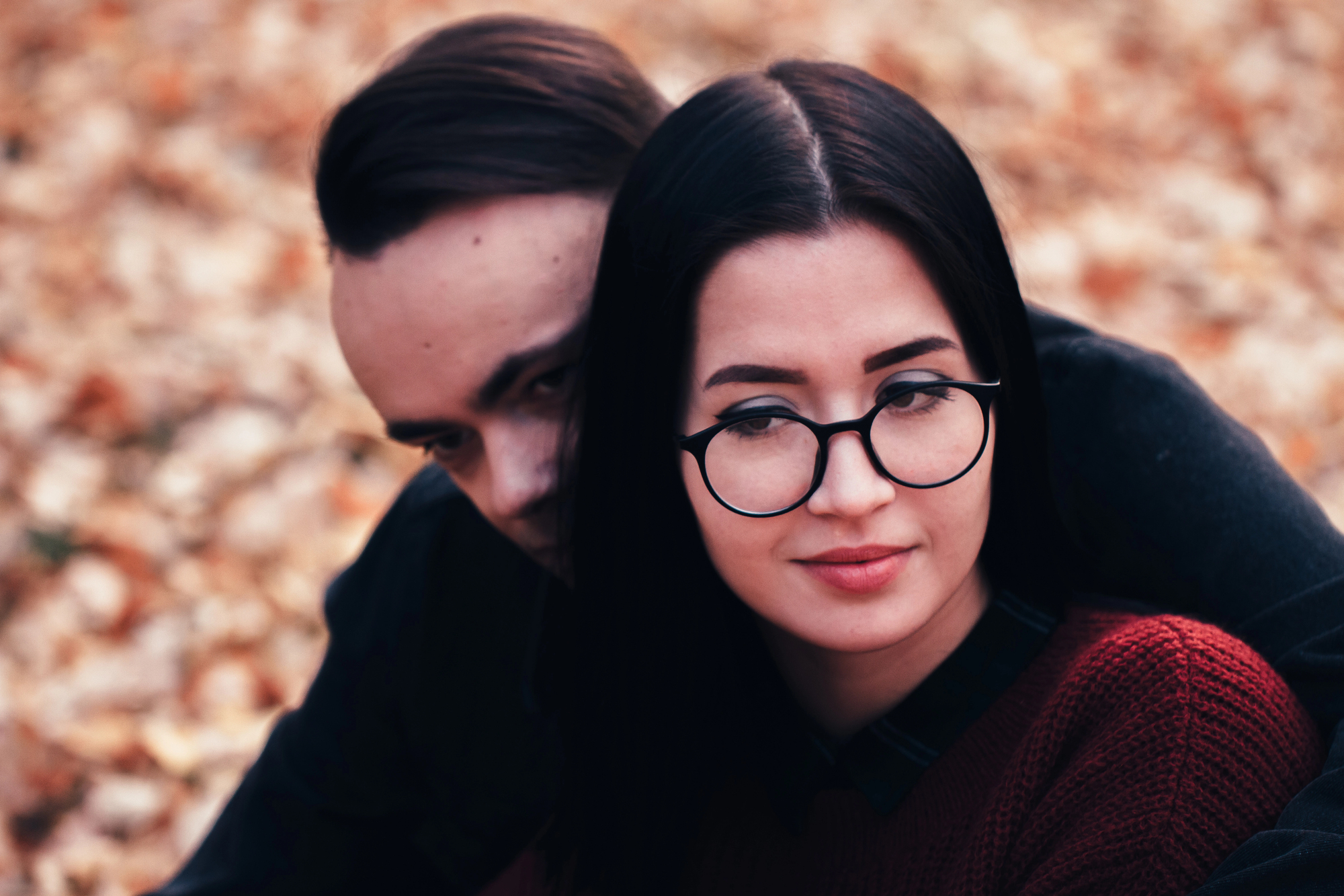 A woman with glasses and dark hair, wearing a red sweater, is sitting in front of a man with dark hair. They are outdoors with autumn leaves on the ground. The man partially hides his face behind her shoulder.