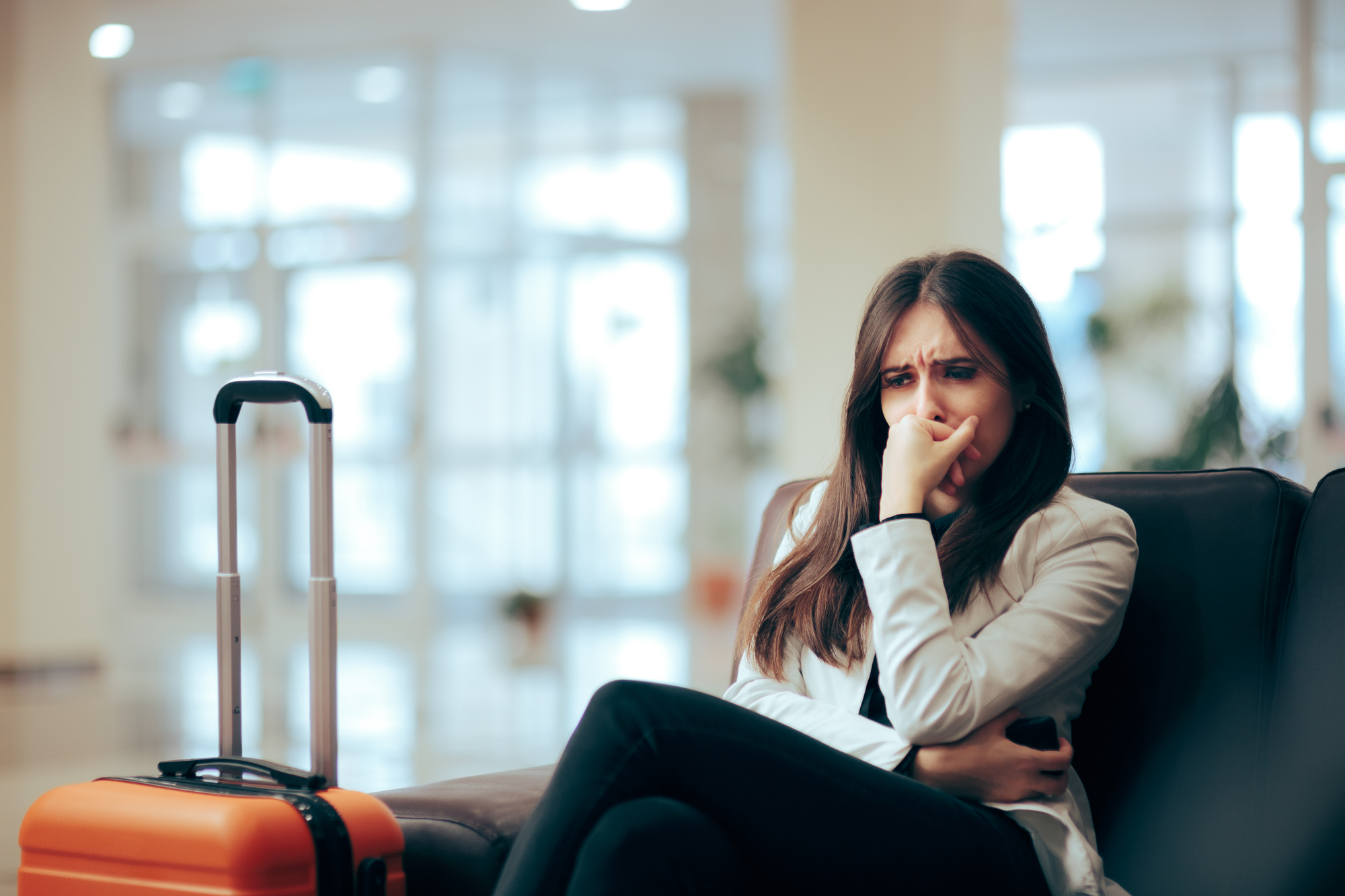 A worried woman sits on a bench in an airport, resting her chin on her hand. An orange suitcase stands beside her. The background is blurred, suggesting a busy terminal with bright lighting.