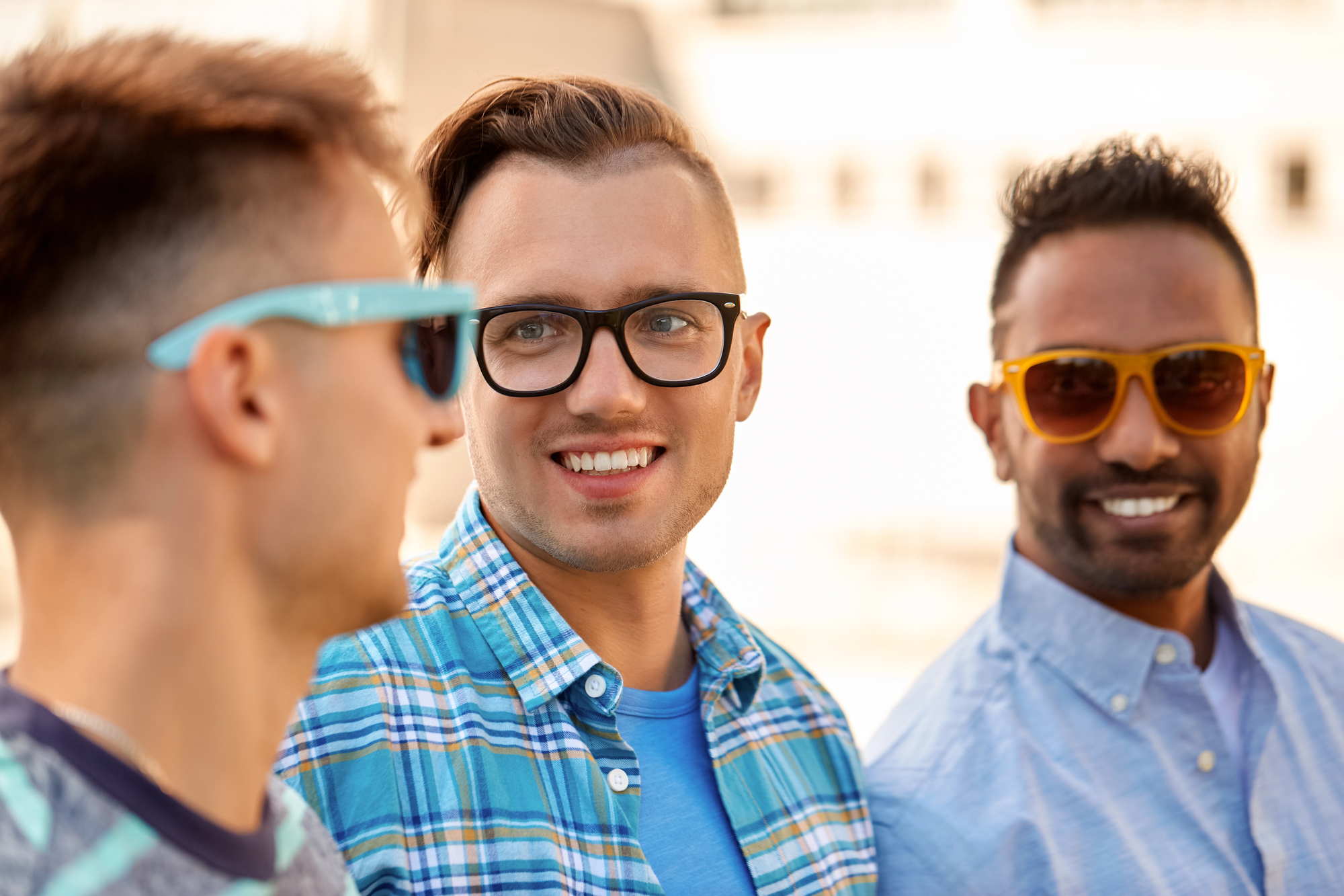 Three men are standing outdoors, smiling and wearing sunglasses. The man in the middle wears glasses and a checkered shirt, the one on the right has a light blue shirt, and the one on the left has blue sunglasses. The background is blurred.