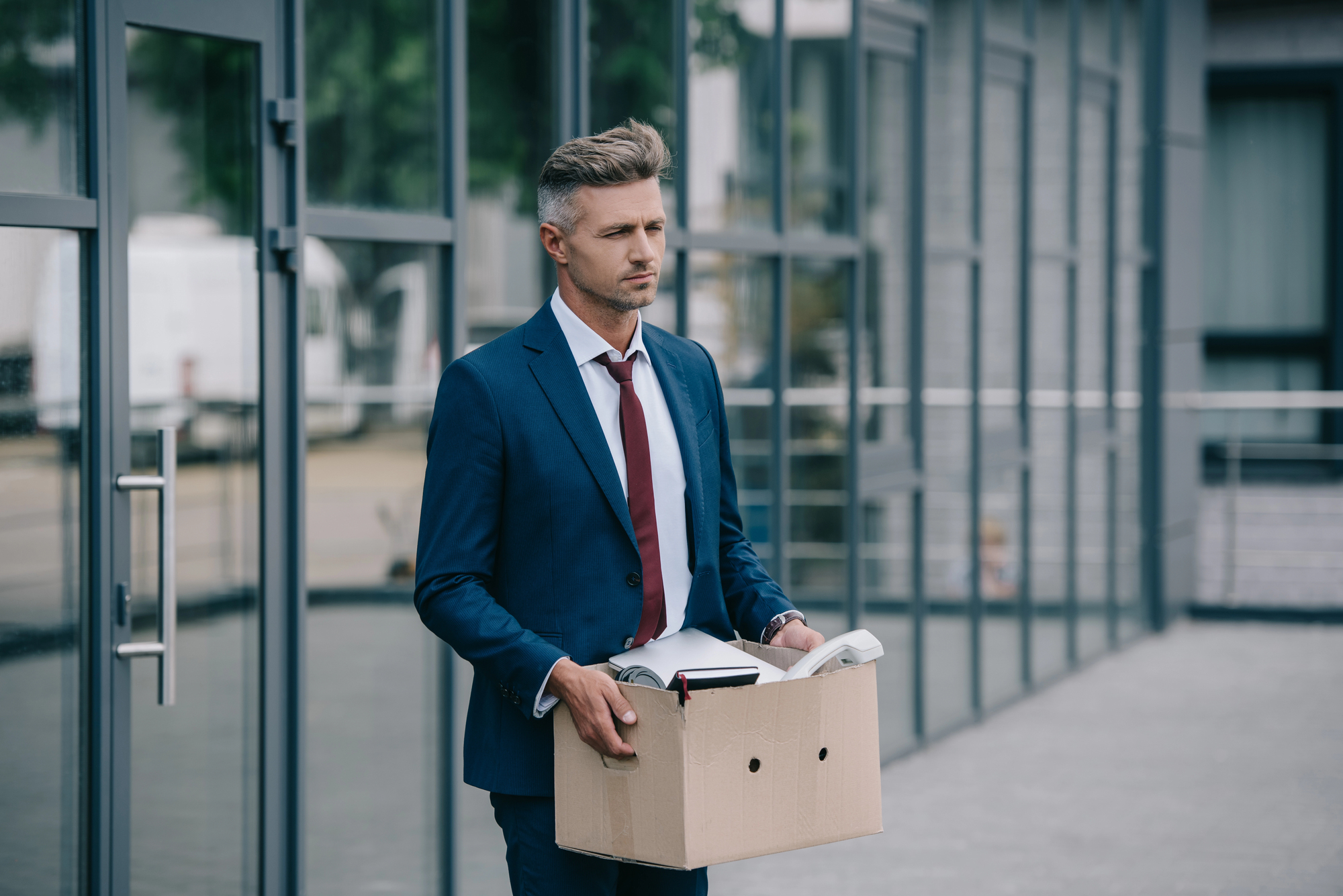 A man in a blue suit carrying a cardboard box, walking outside a modern glass building. He looks pensive, and the box contains office supplies, suggesting he might be leaving an office job.