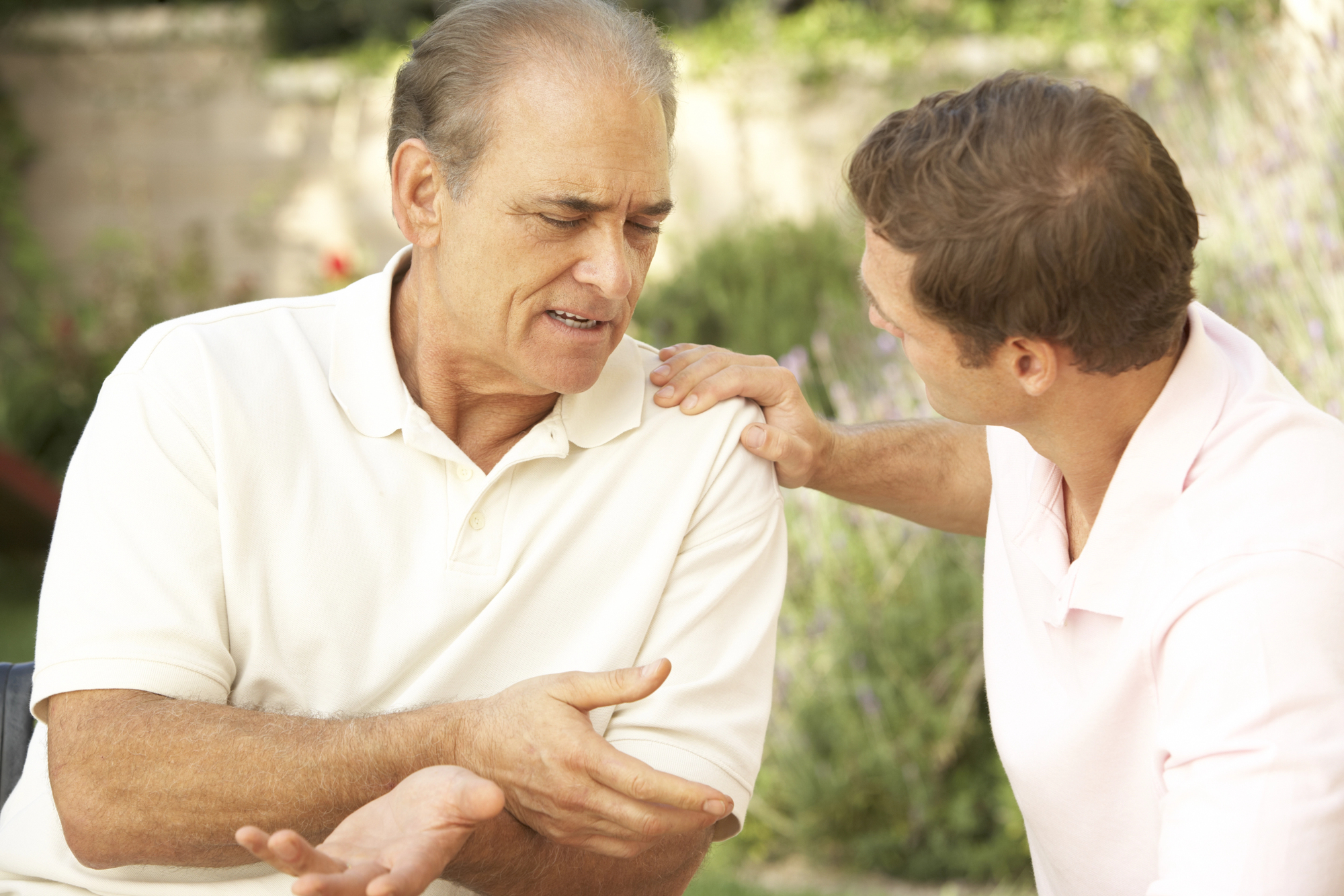 A younger man is sitting outside, gently placing his hand on the shoulder of an older man with graying hair. The older man appears to be speaking, with a thoughtful expression, while they both sit in a garden setting.